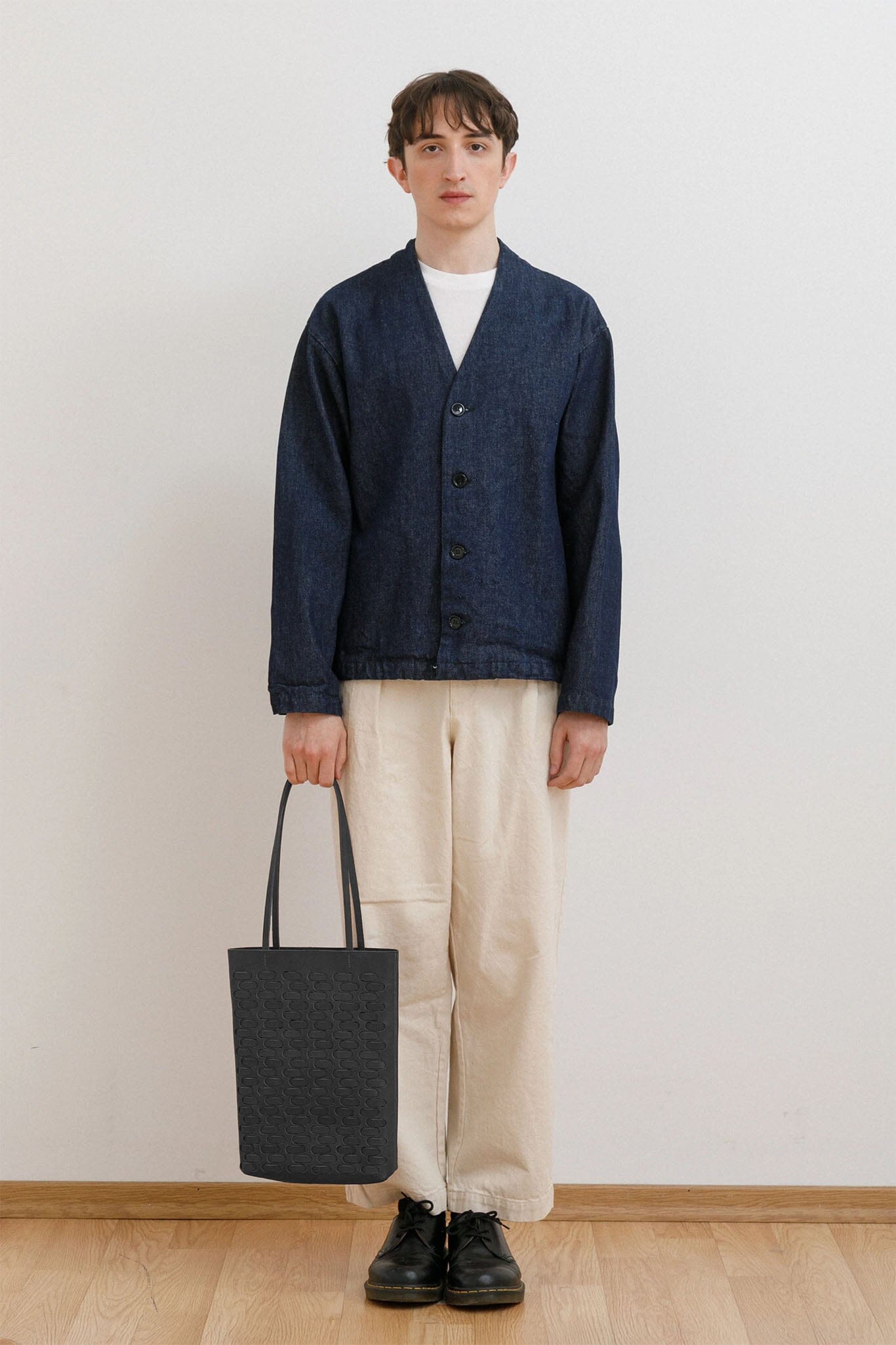 Man wearing a navy cardigan, white shirt, beige pants, and black shoes, holding a black woven tote bag against a plain background.