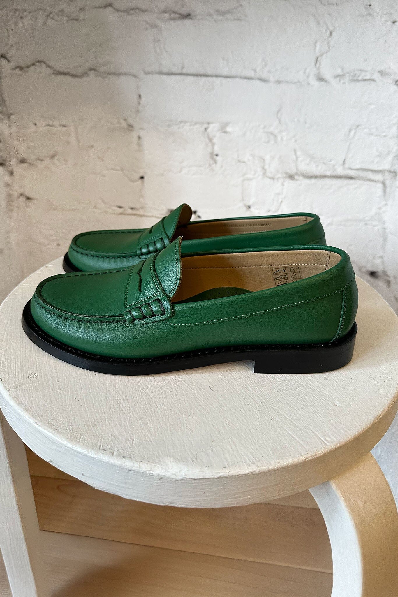 Green leather loafers on a round wooden table with a white brick wall background