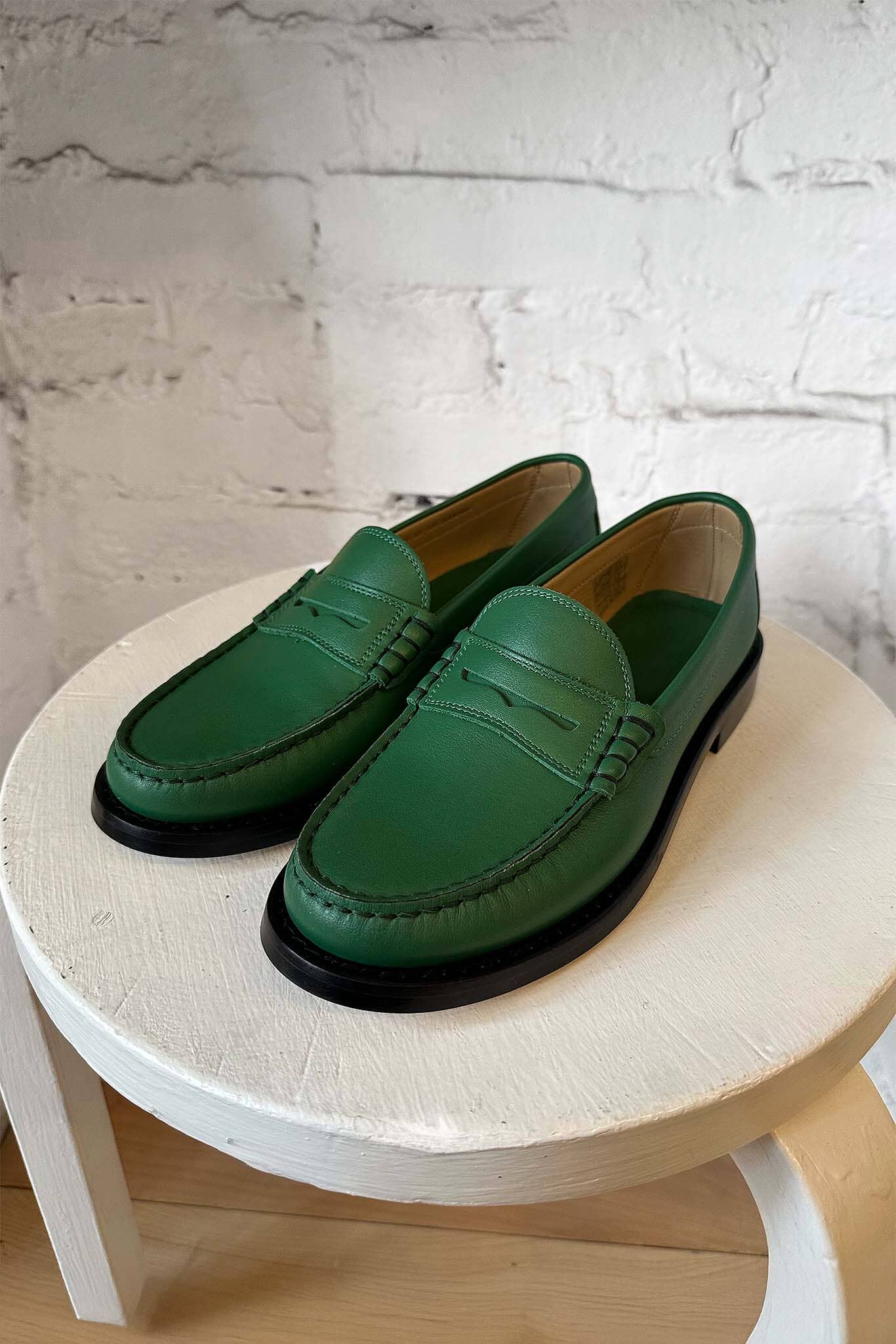 Green leather loafers on a round wooden stool against a white brick wall.
