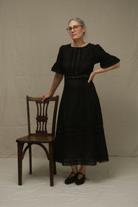 A woman wearing a black midi dress with puff sleeves and a lace-trimmed neckline stands next to a wooden chair against a neutral beige backdrop.