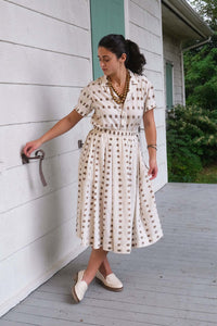 A woman wearing a cream-colored midi dress with a brown geometric pattern stands on a gray wooden porch next to a white house wall.