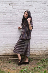 A woman wearing a matching plaid and striped long-sleeve top and midi skirt set, posing against a white painted brick wall outdoors.
