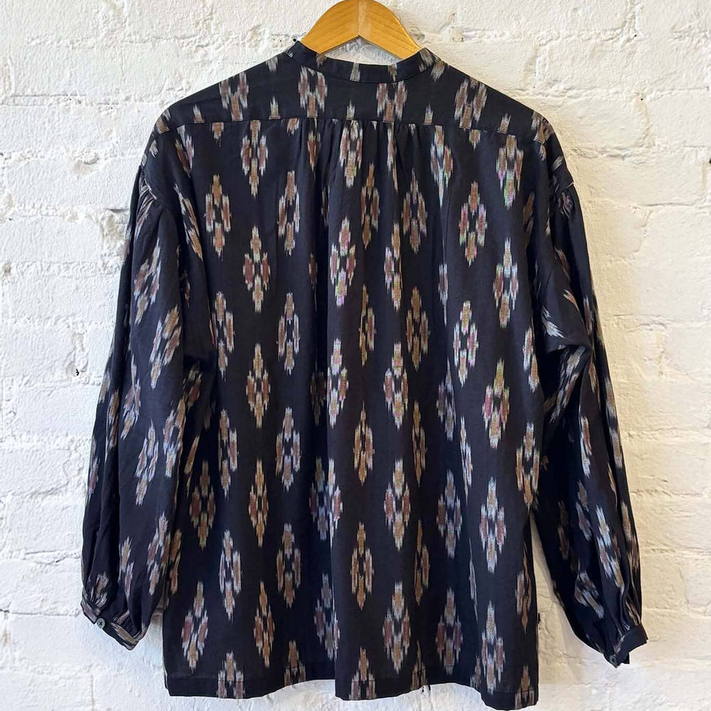The back view of a black patterned blouse on a wooden hanger against a white brick wall.