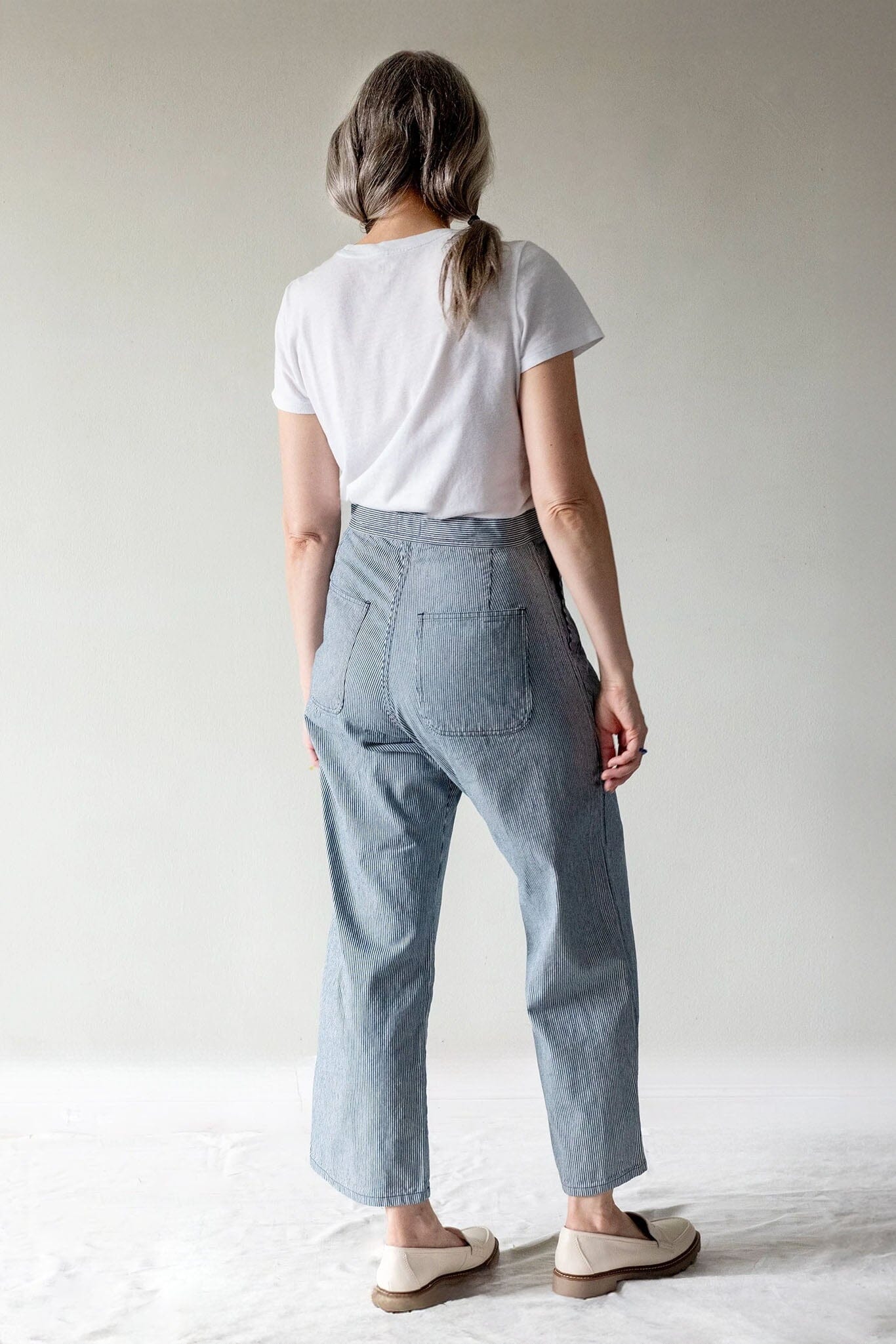 A woman shown from behind wearing light blue wide-leg cropped jeans, a white t-shirt, and cream-colored slip-on shoes against a neutral studio backdrop.
