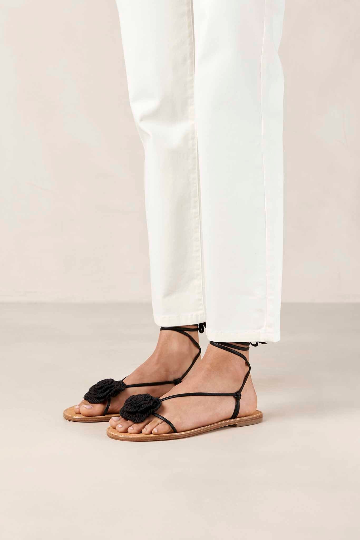 A pair of black leather ALOHAS Jakara sandals with crocheted flowers on the straps, displayed on a person wearing white pants.