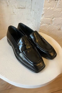 Pair of black loafers on a white pedestal with a light-colored brick wall background