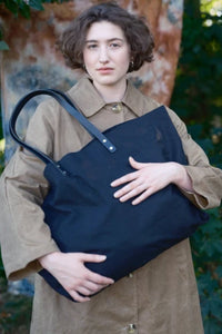 A woman wearing a brown trench coat holds a large black canvas tote bag with dark leather shoulder straps in an outdoor setting.