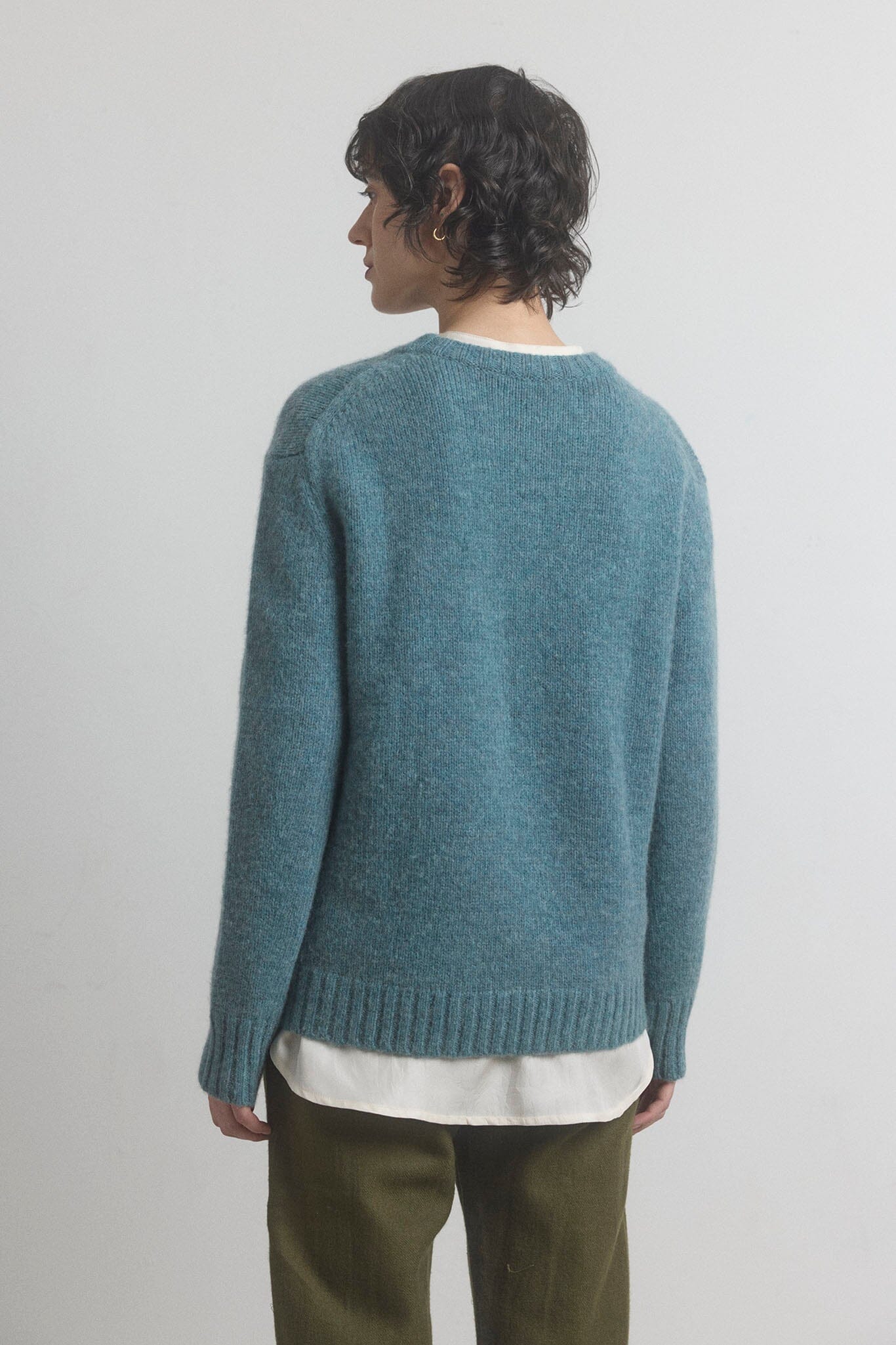 Woman facing the back wearing a blue merino knitted sweater with a plain background