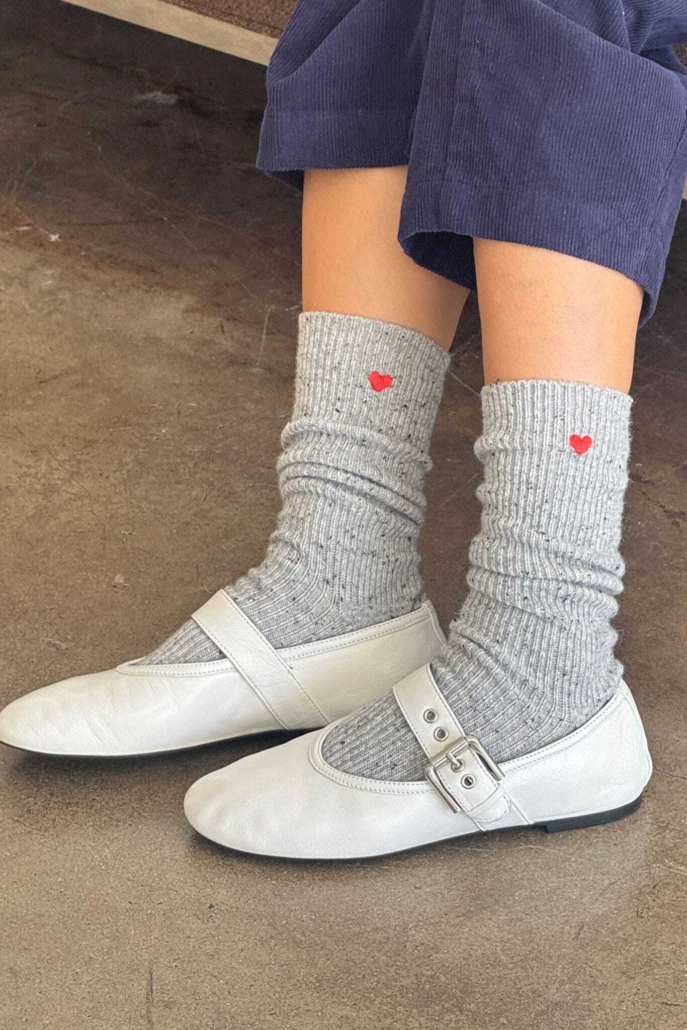 Gray socks with red hearts worn by a person in white ballet flats on a concrete floor.