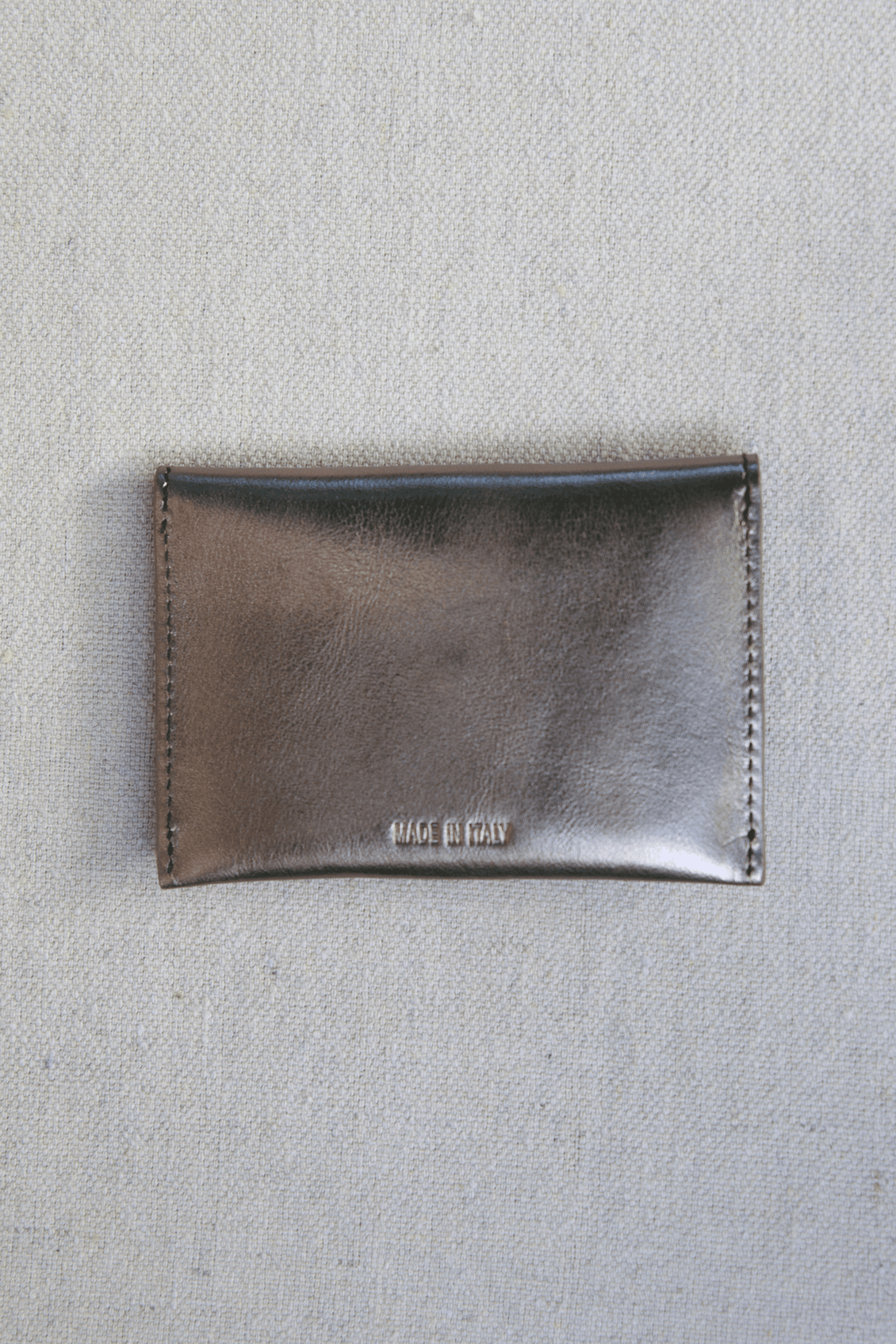 A small metallic bronze leather card holder or wallet with visible stitching and the debossed brand name 'MADE IN ITALY' on the front, placed on a textured light gray fabric background.