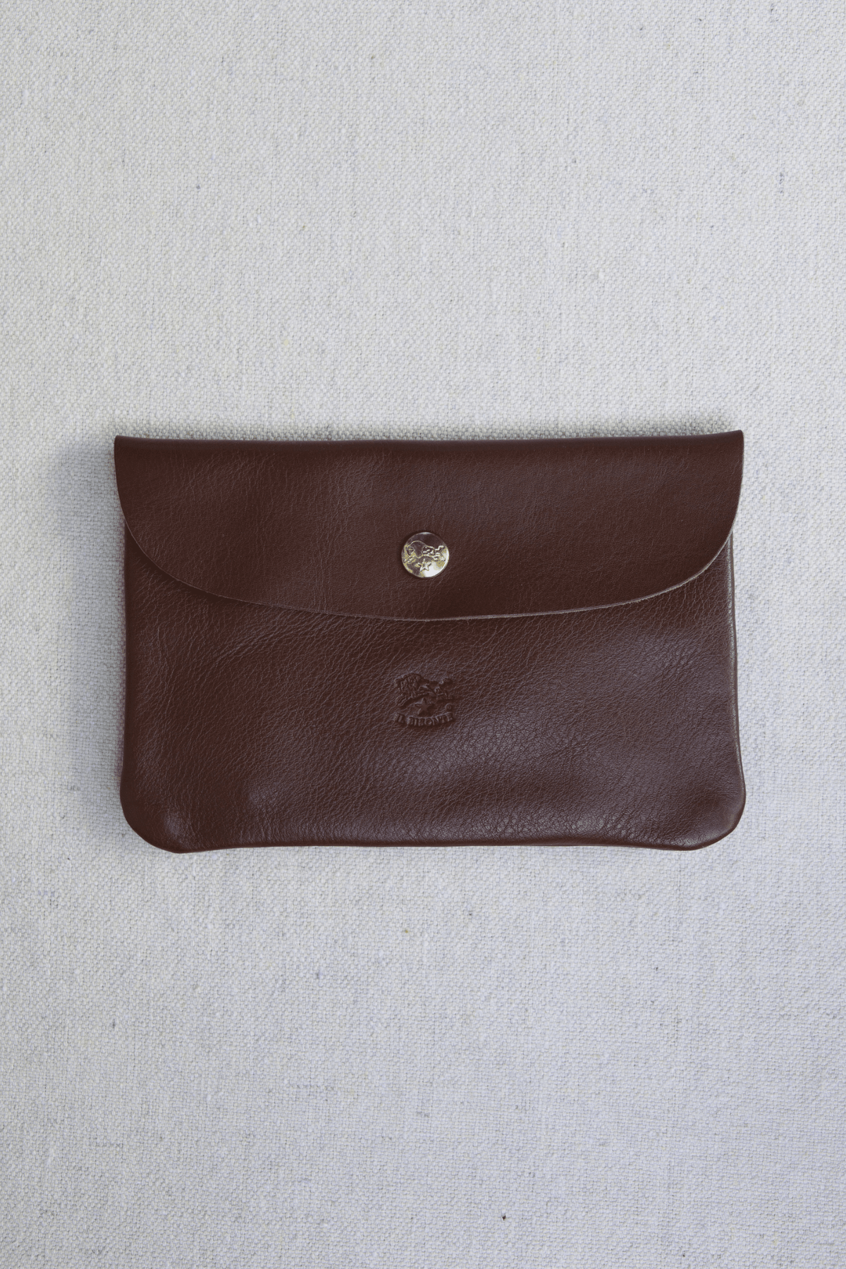 A dark brown leather envelope-style pouch with a gold snap button closure, laid flat on a light beige textured fabric background.