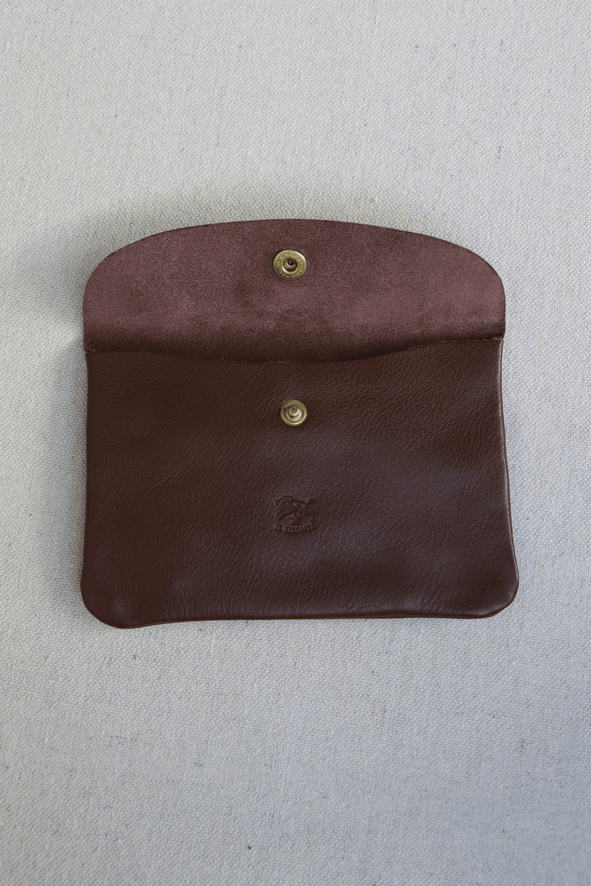 A dark brown leather pouch with a curved top flap and two brass snap buttons, laid flat on a light gray textured background.
