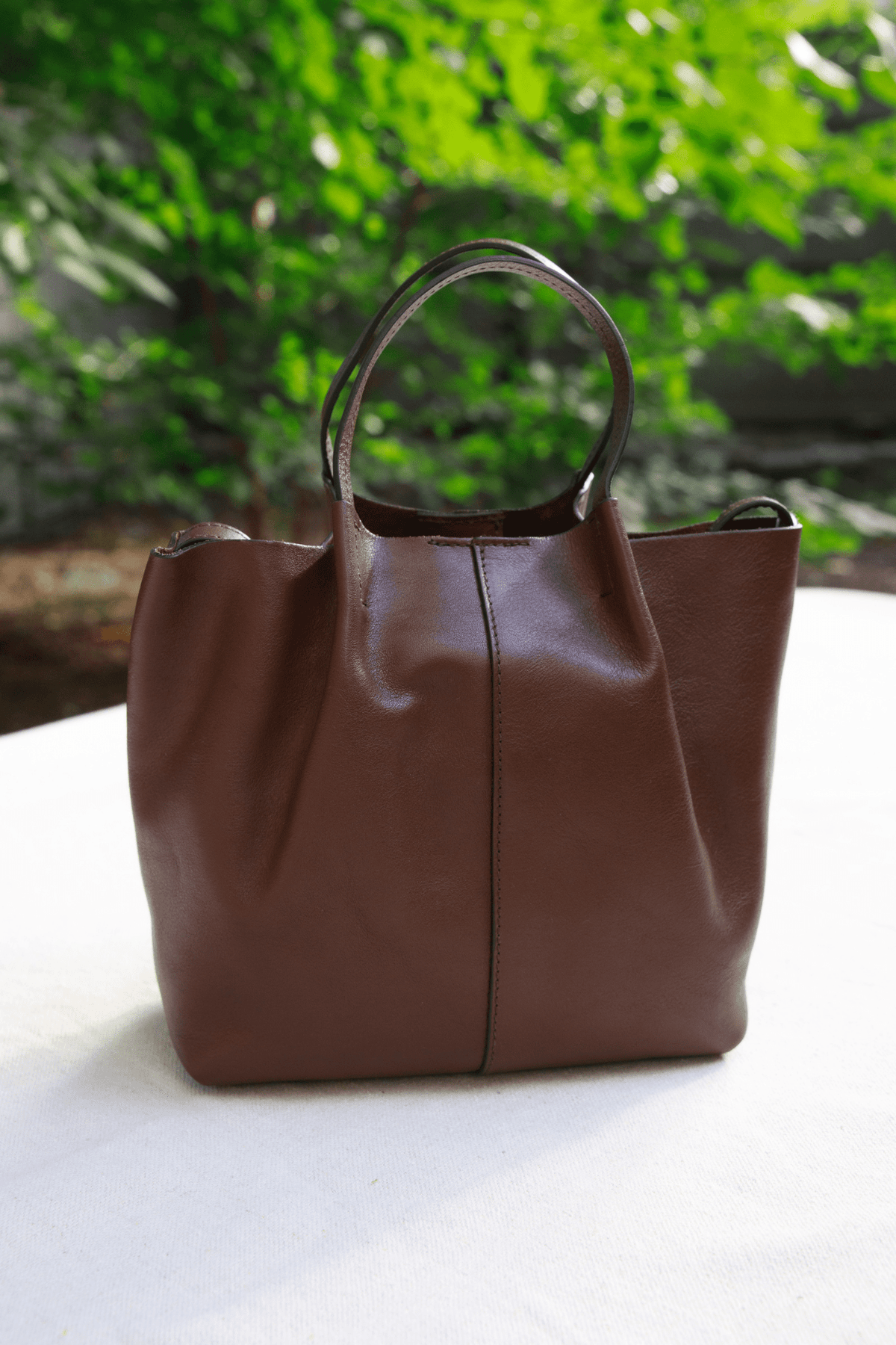 A brown leather tote bag with a top handle, standing on a white surface with a blurred green garden background.
