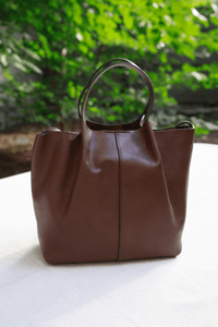 A brown leather tote bag with a top handle, standing on a white surface with a blurred green garden background.