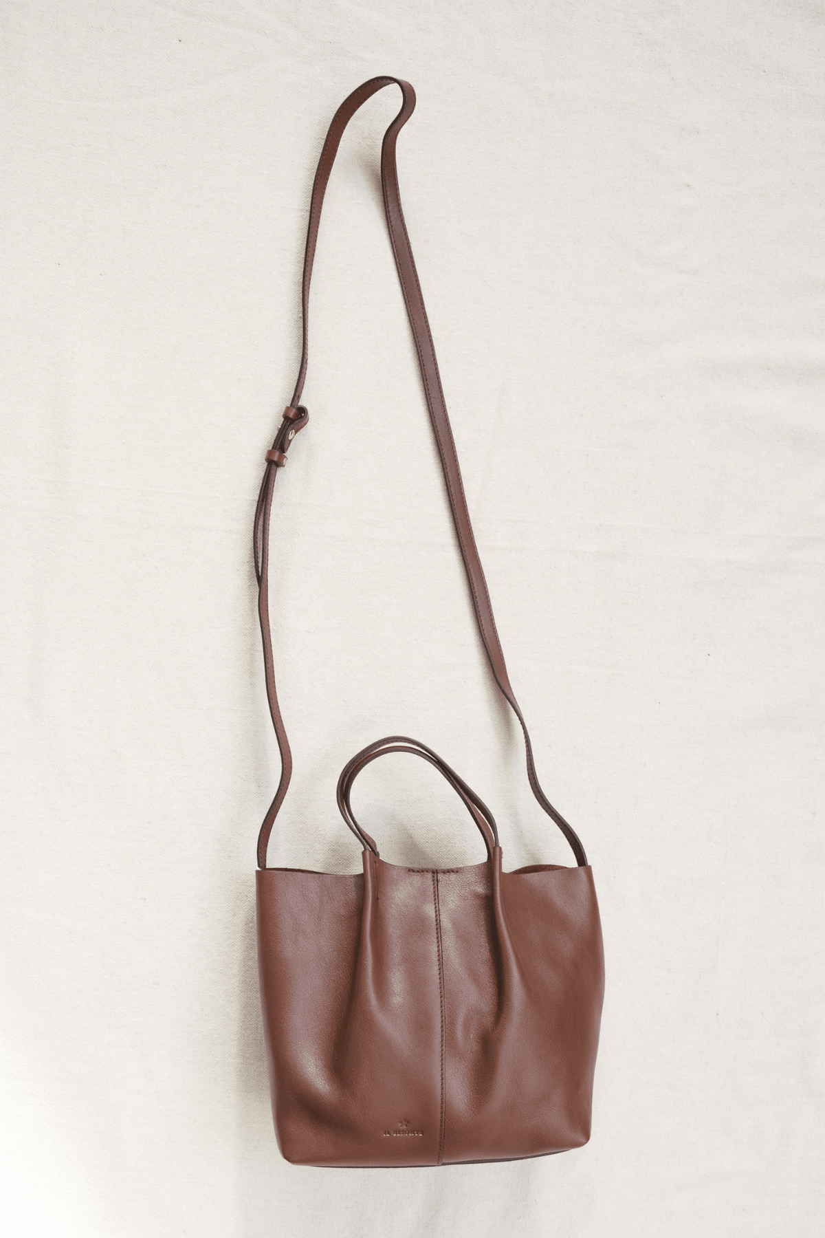 A brown leather shoulder bag with a long adjustable strap and top handles, laid flat on a light off-white fabric background.