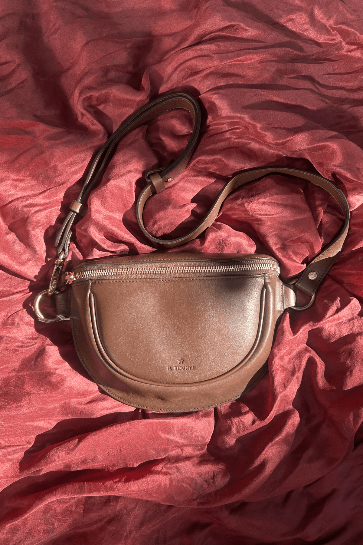 A brown leather crossbody bag with a curved front pocket and adjustable shoulder strap, laid flat on a draped dusty rose fabric background.