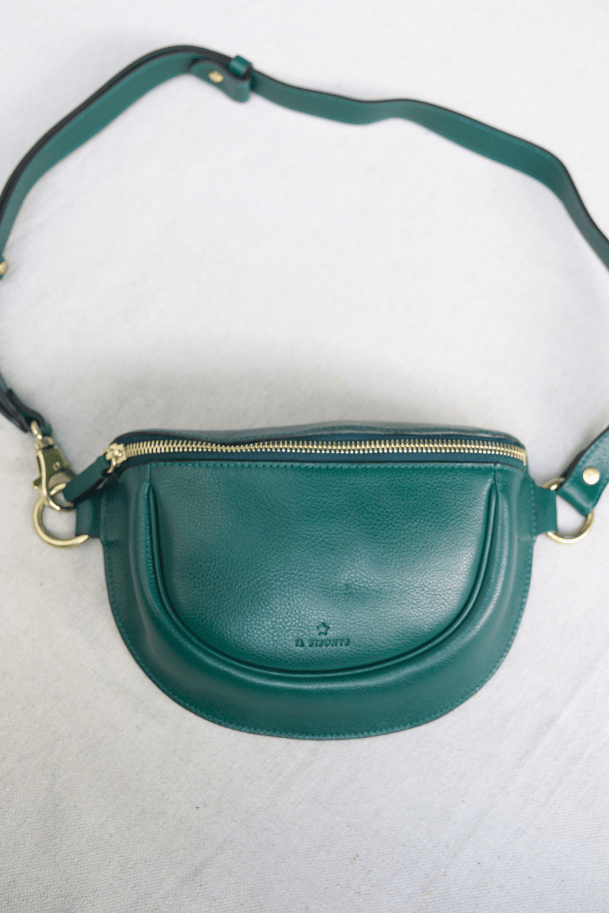 A green leather crescent-shaped crossbody bag with a gold zipper and gold-tone hardware, laid flat on a light gray fabric background.
