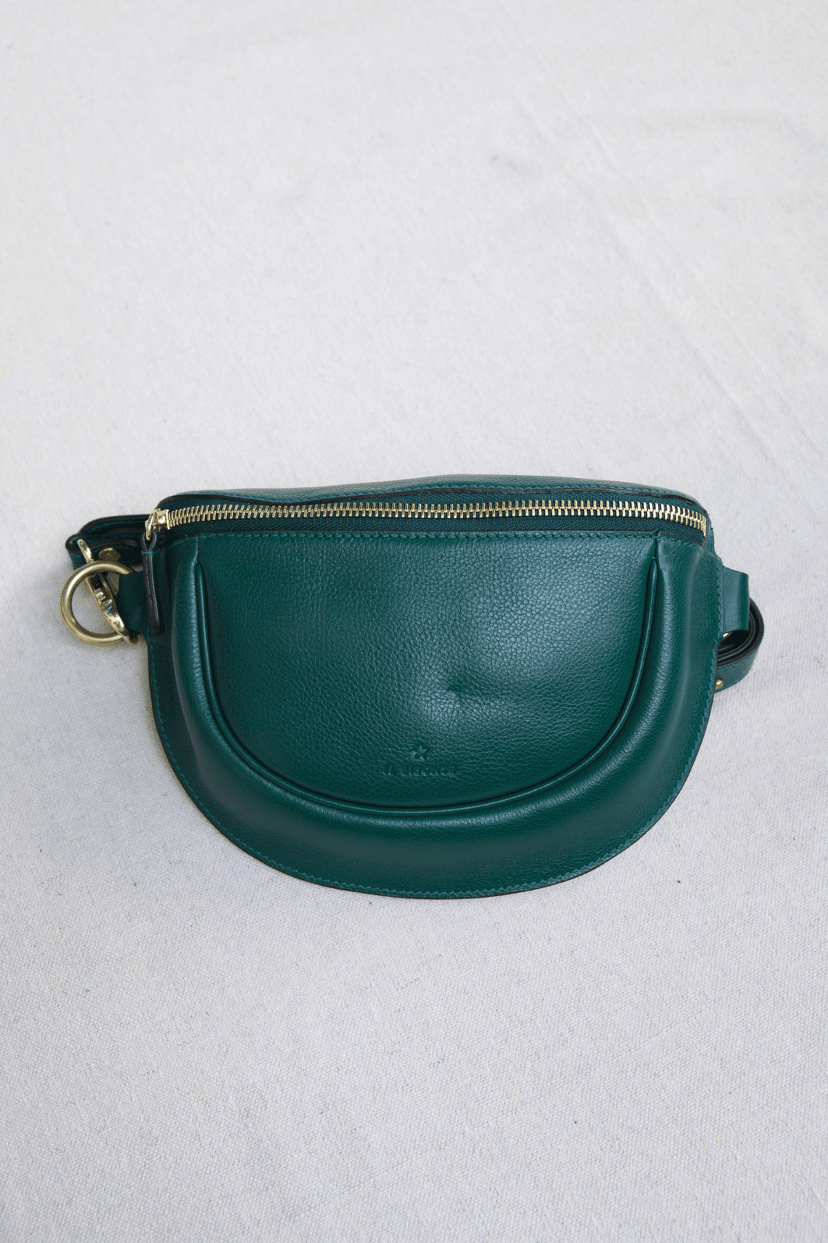 A dark green leather fanny pack with a gold-tone zipper and hardware, photographed flat on a light gray textured fabric background.