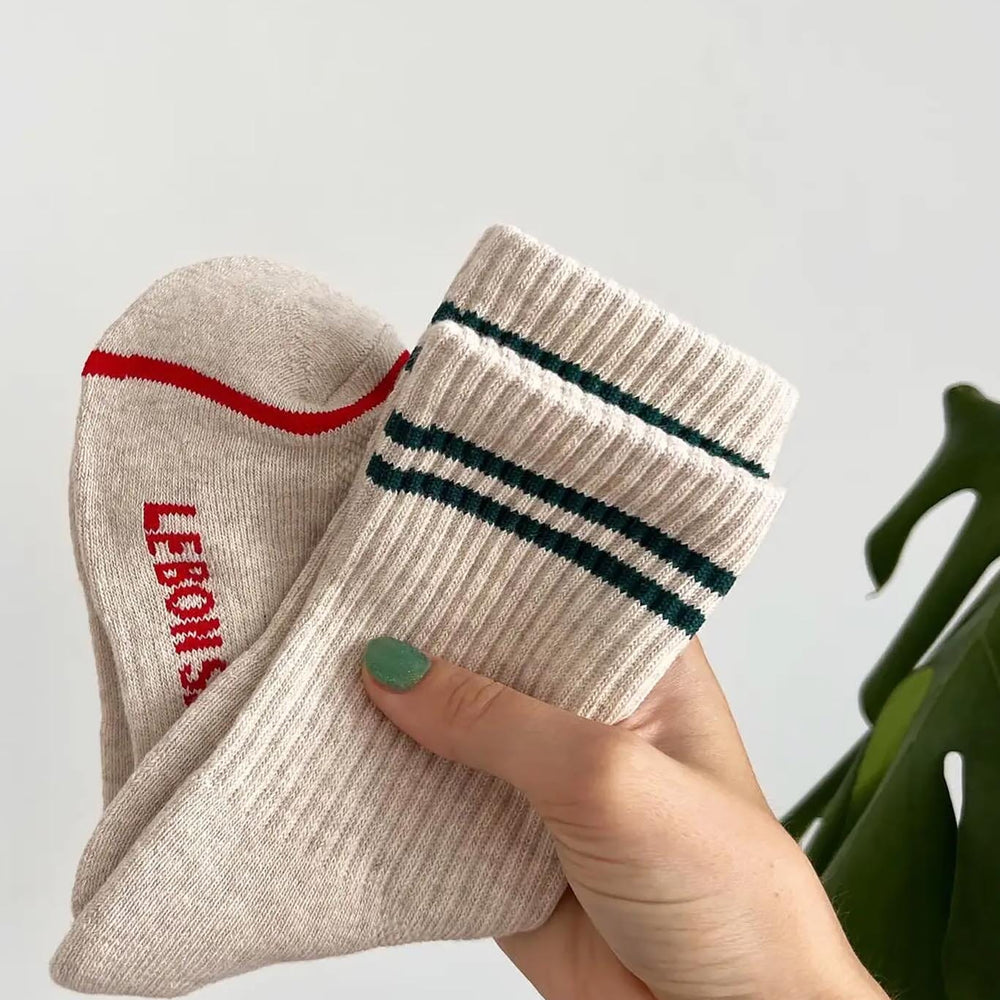 A person holding a pair of oatmeal-colored boyfriend socks with red stripes and the brand name 'LE BON SHOPPE' visible on the foot area.