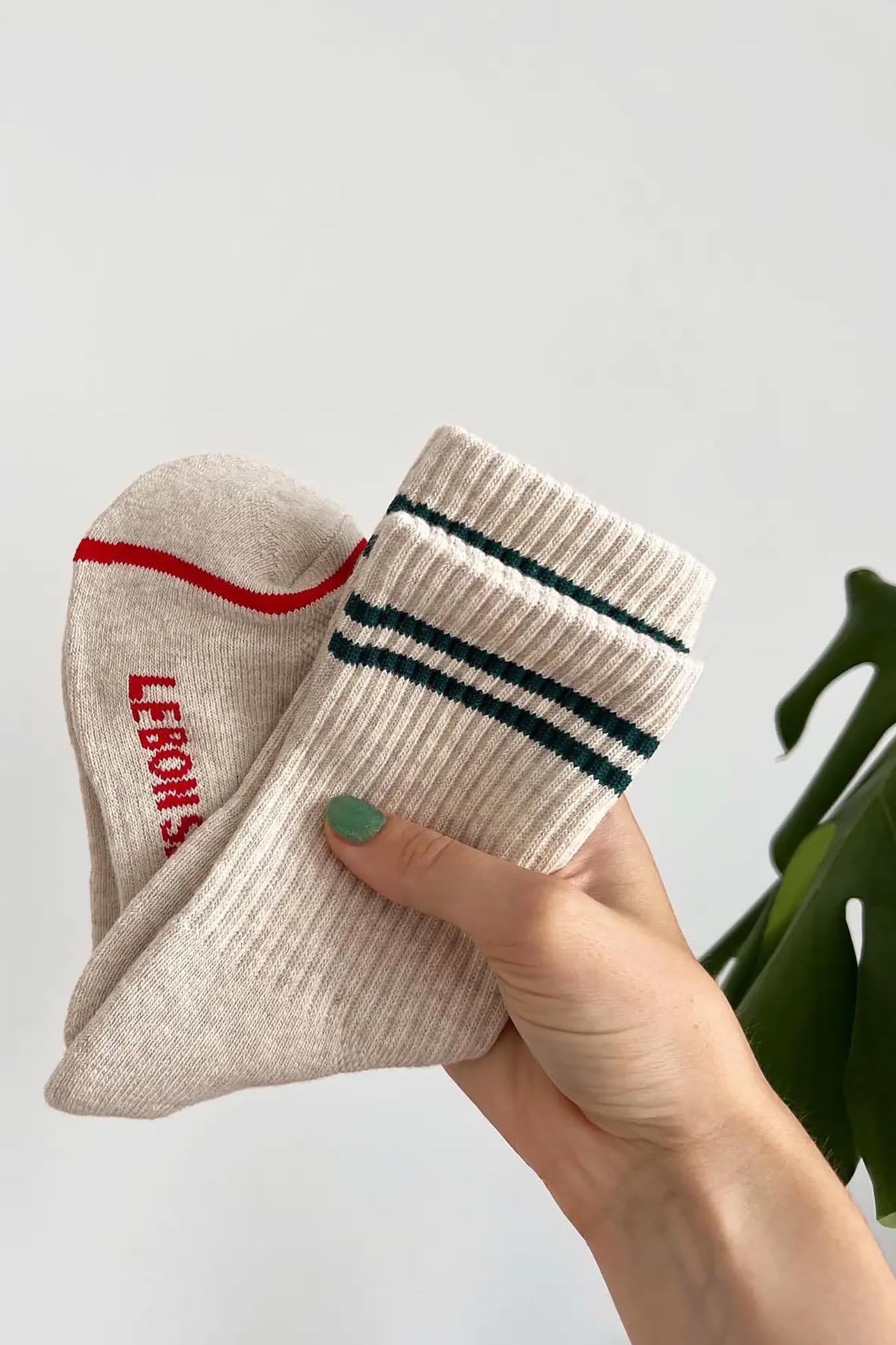 A person holding a pair of oatmeal-colored boyfriend socks with red stripes and the brand name 'LE BON SHOPPE' visible on the foot area.