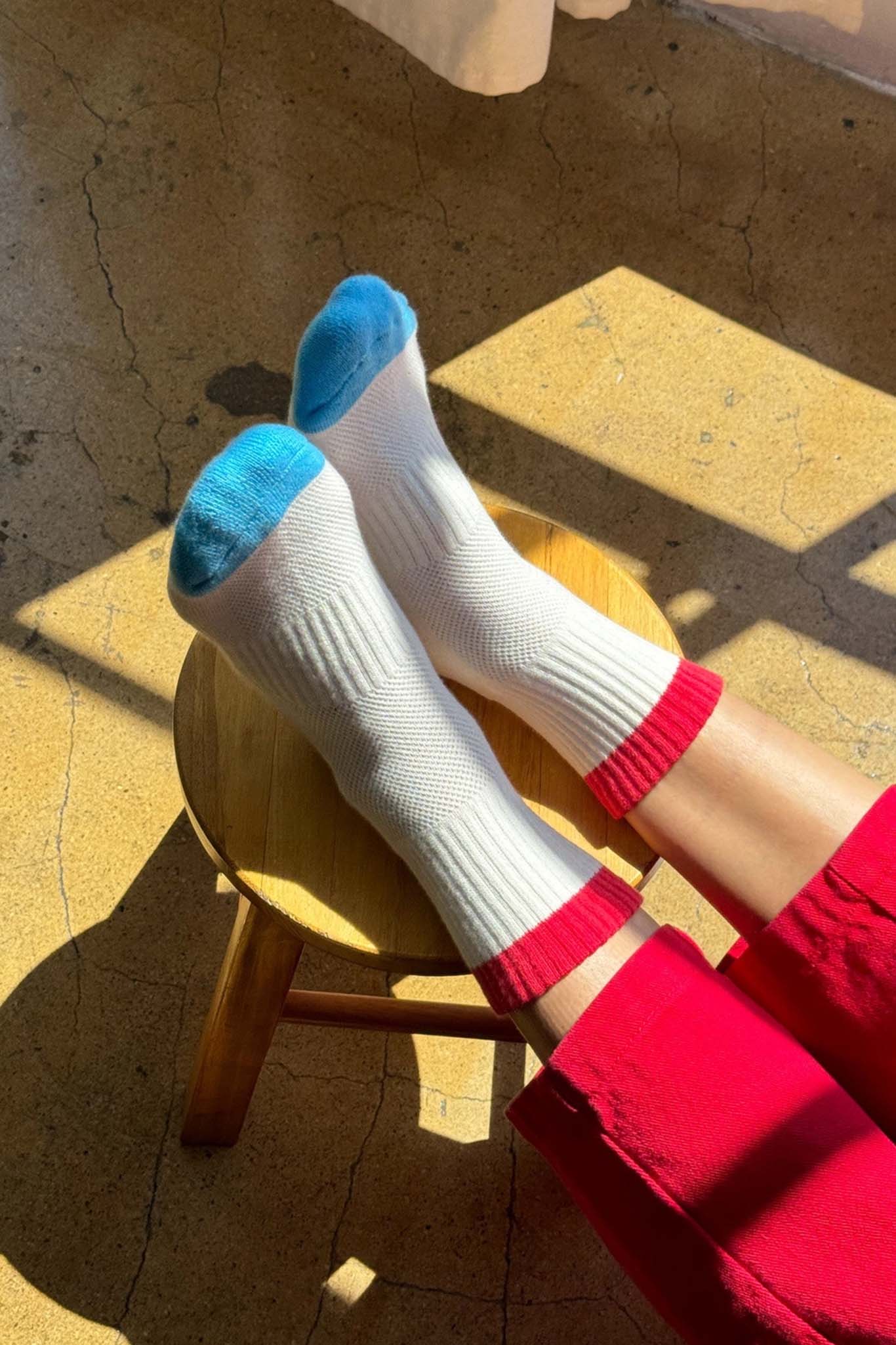 White socks with blue toes and red cuffs on a wooden stool with a sunlit floor.