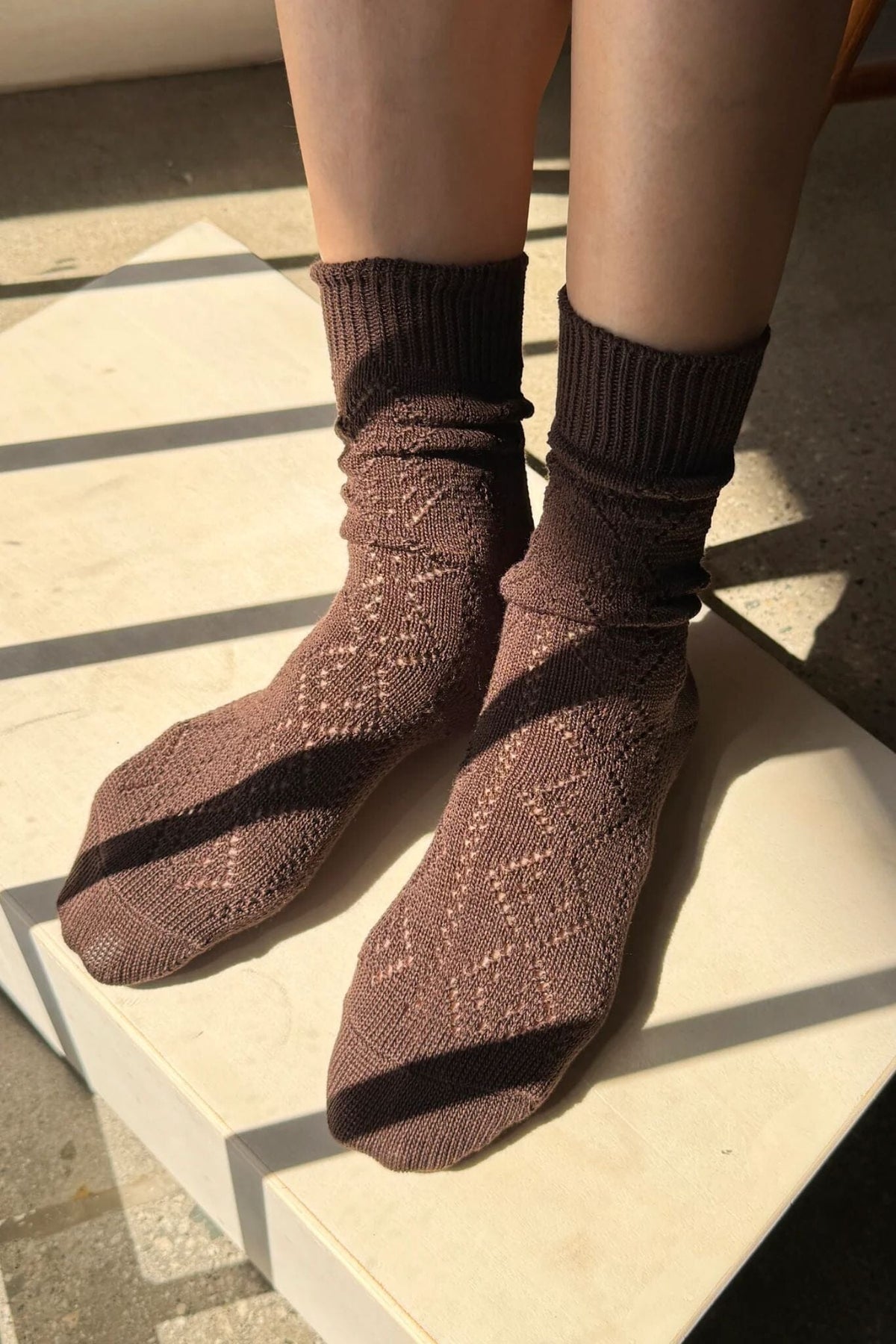 A close-up of a person's feet wearing brown knit socks with a textured openwork pattern and ribbed cuffs, standing on a light stone surface in bright sunlight.