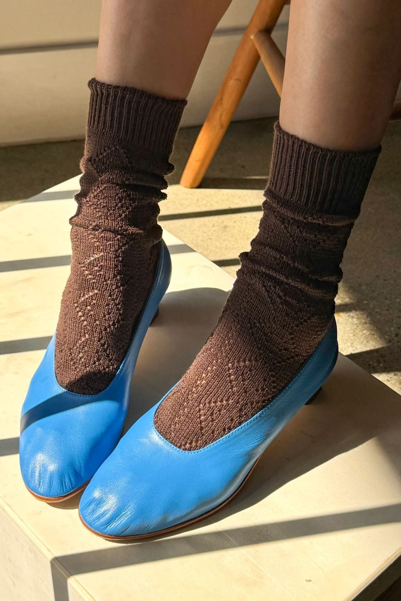 A person wearing bright blue slip-on shoes with brown ribbed socks, photographed in warm sunlight on a light wooden surface.