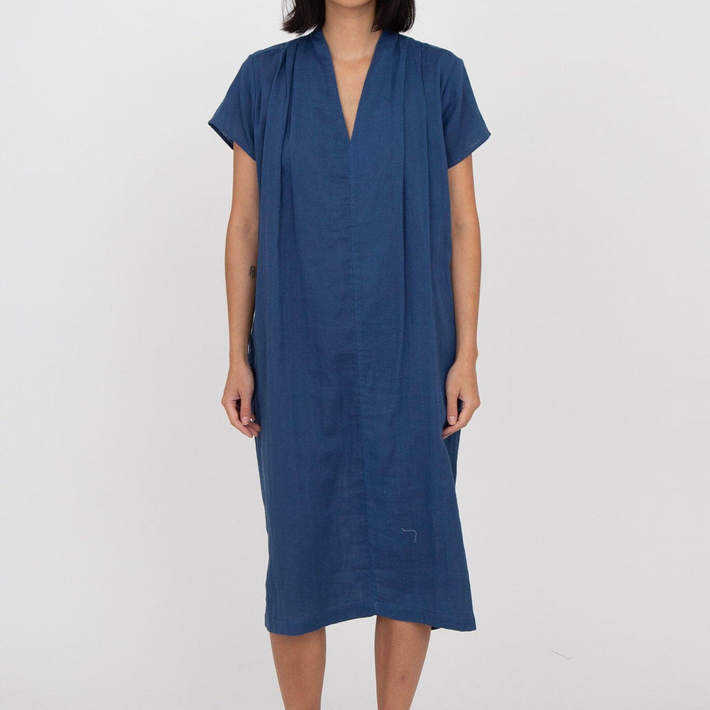 A woman standing in front of a white background wearing a blue v-neck dress with cap sleeves and side seam pockets, reaching just below the knees.