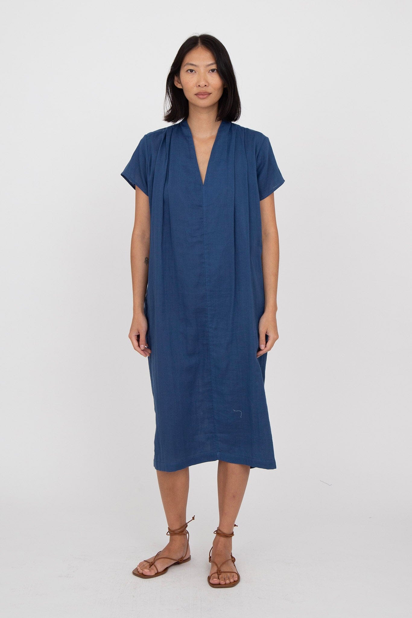 A woman standing in front of a white background wearing a blue v-neck dress with cap sleeves and side seam pockets, reaching just below the knees.