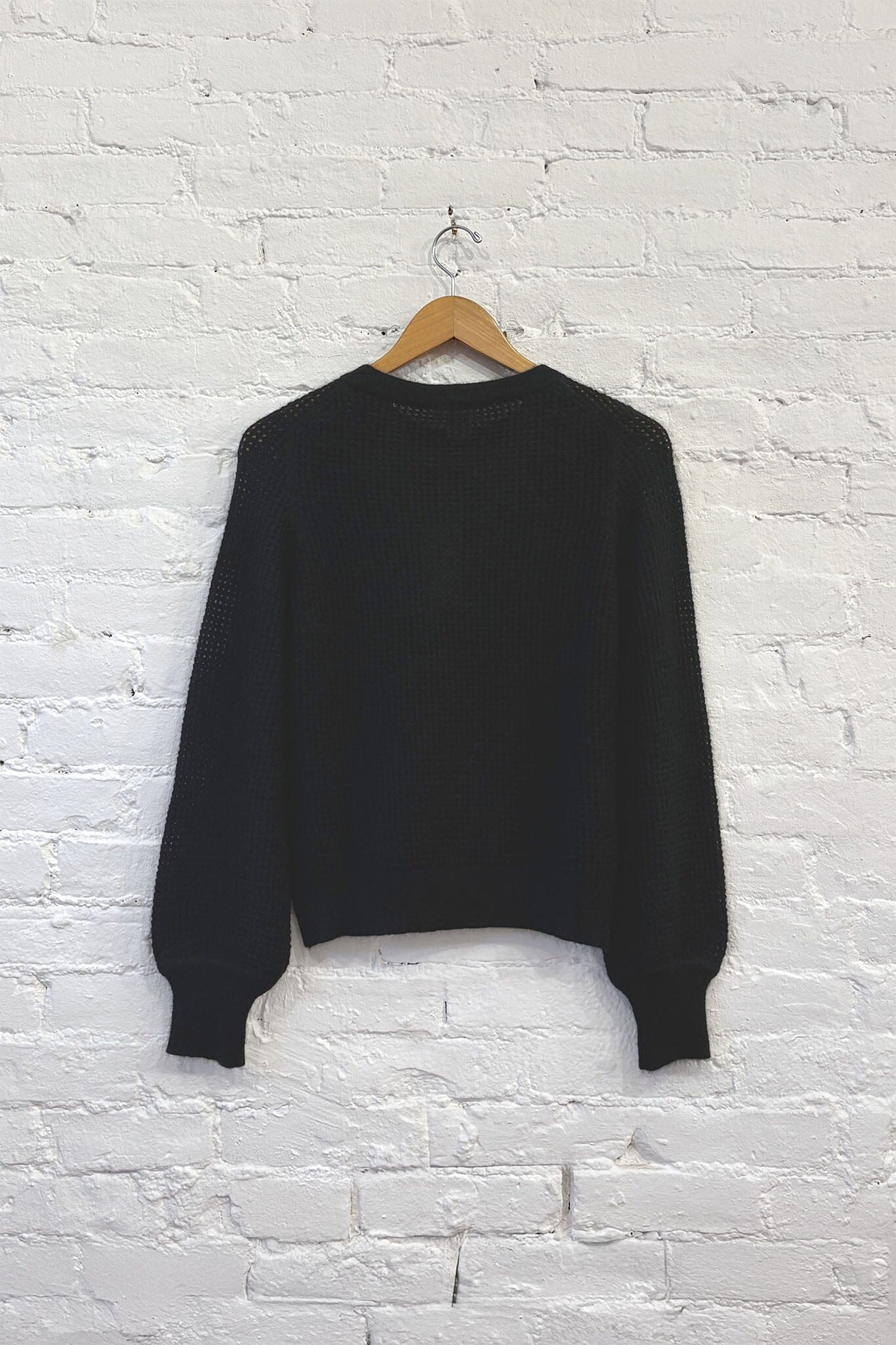 The back of a black knit cashmere sweater on a wooden hanger against a white brick wall
