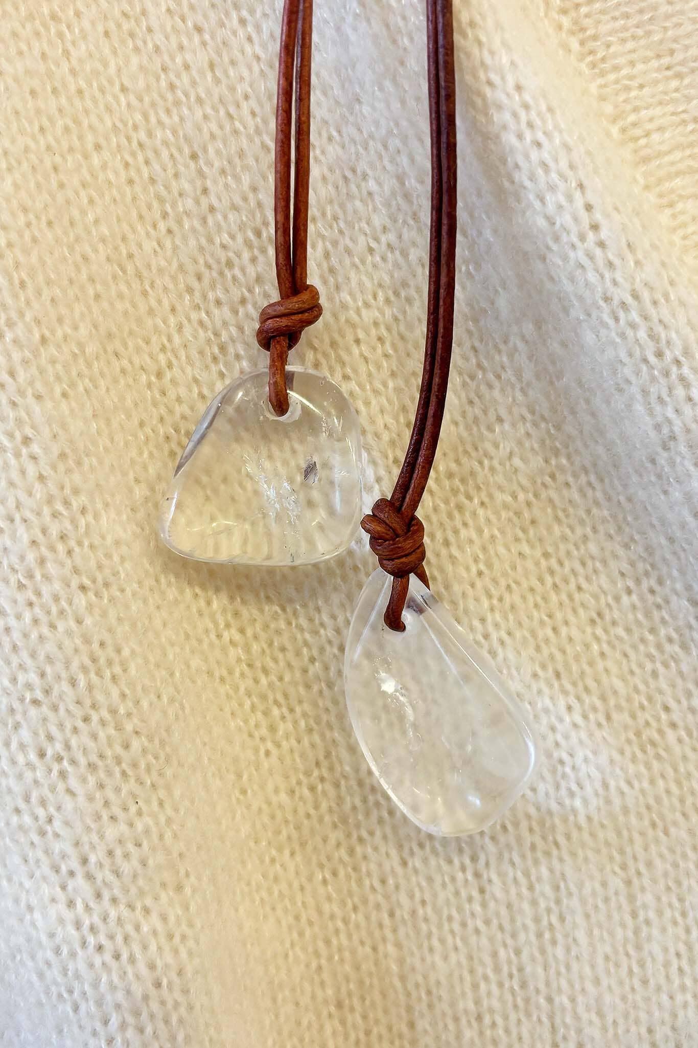 Clear teardrop-shaped pendant on a brown leather cord against a beige fabric background