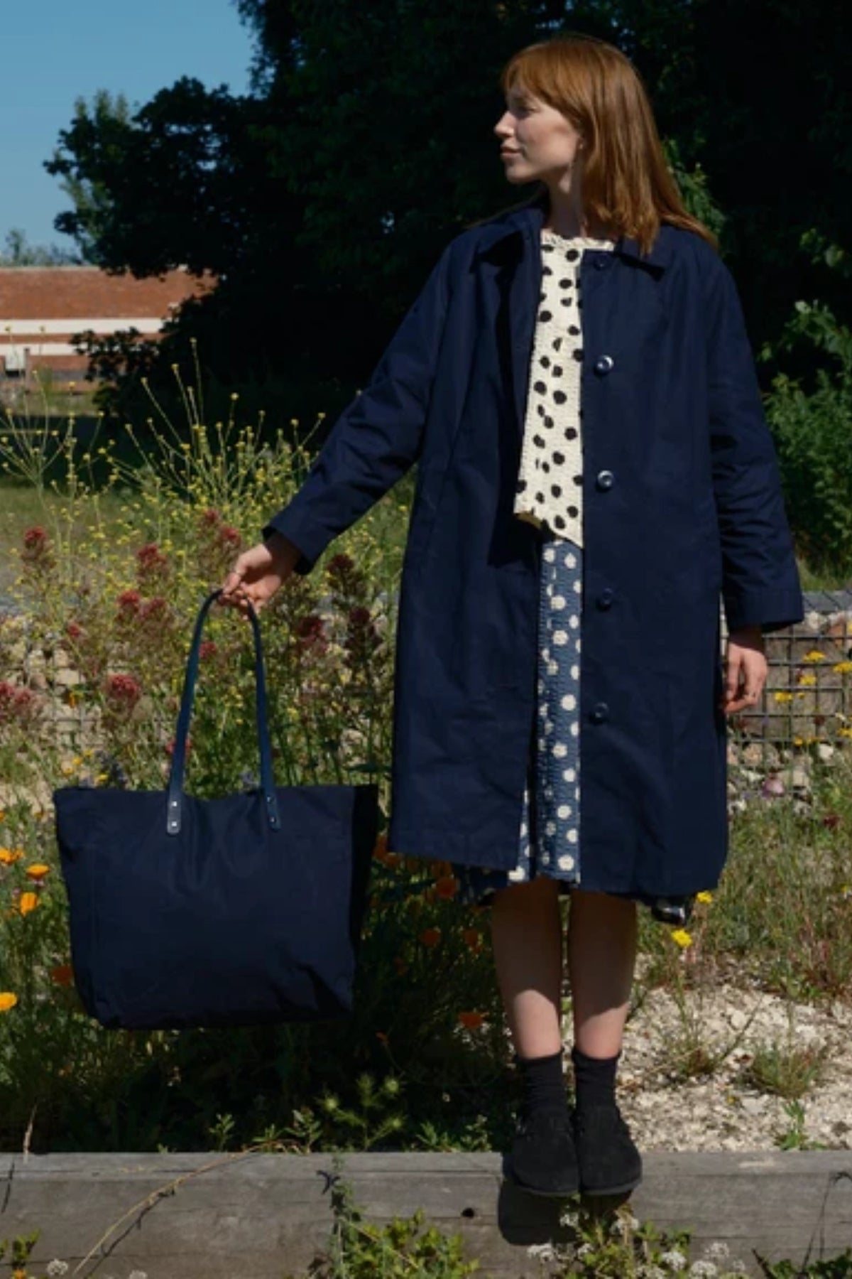 A woman standing outdoors in a garden wearing a long navy coat over a cream dress with white polka dots, holding a navy tote bag.