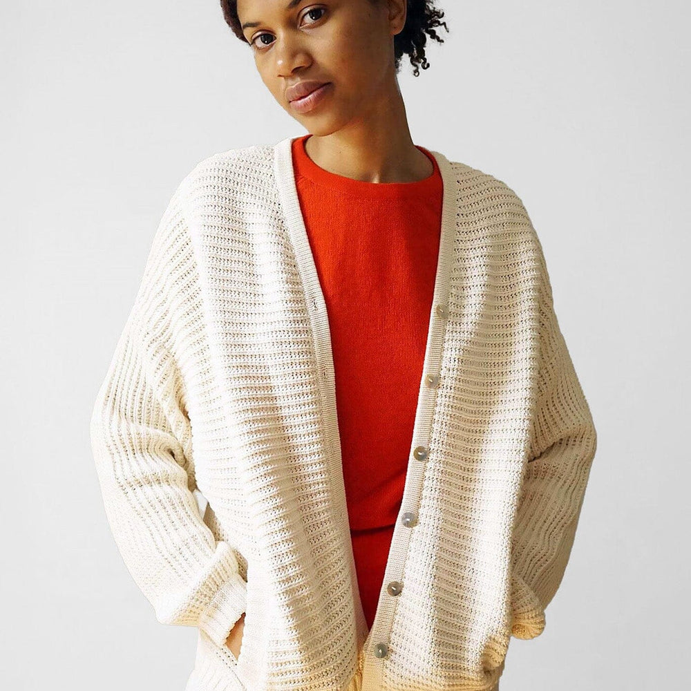 A woman wears a red shirt beneath a cozy long sleeve, cream and cotton sweater. Designed by M.Patmos in Brooklyn, New York.