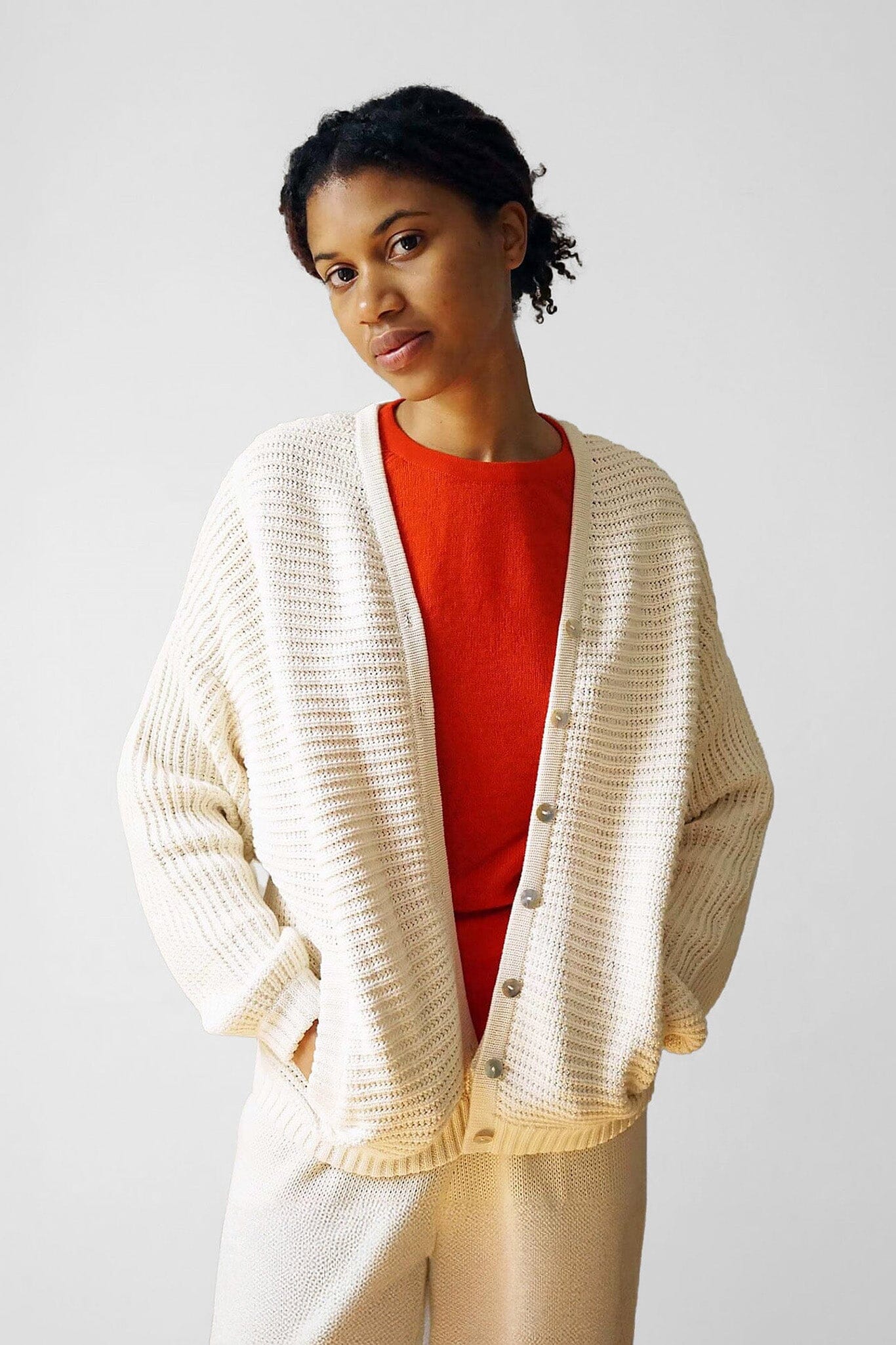 A woman wears a red shirt beneath a cozy long sleeve, cream and cotton sweater. Designed by M.Patmos in Brooklyn, New York.