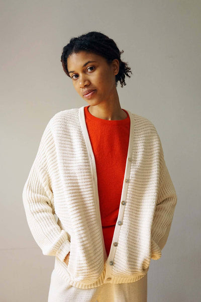 A woman wears a red shirt beneath a cozy long sleeve, cream and cotton sweater. Designed by M.Patmos in Brooklyn, New York.