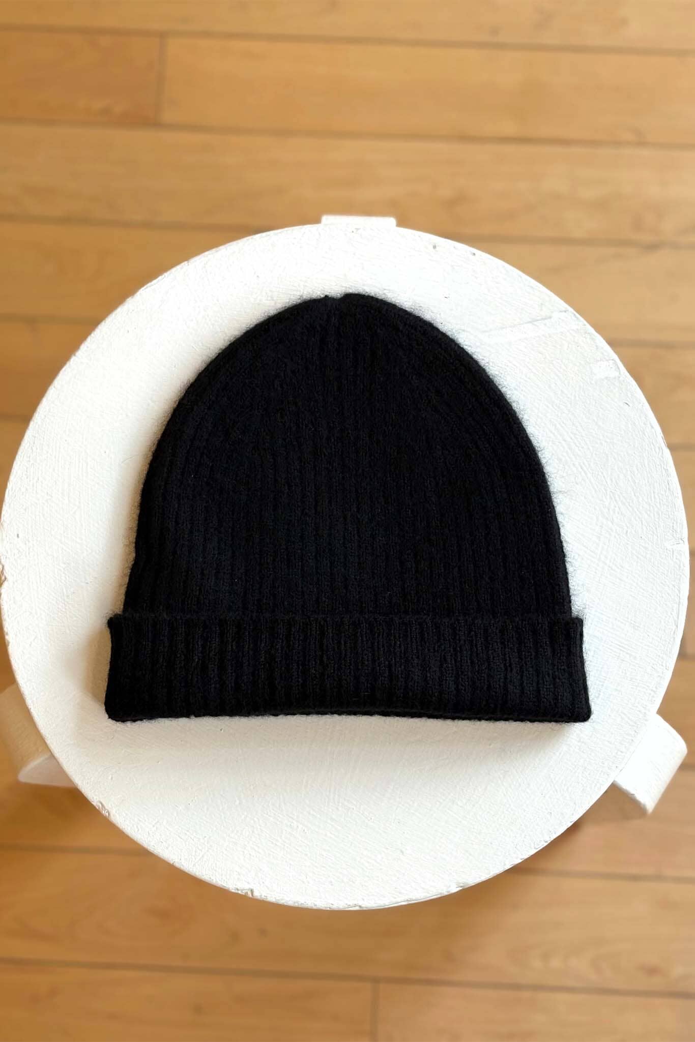 Black knit beanie on a white circular cushion with a wooden floor background