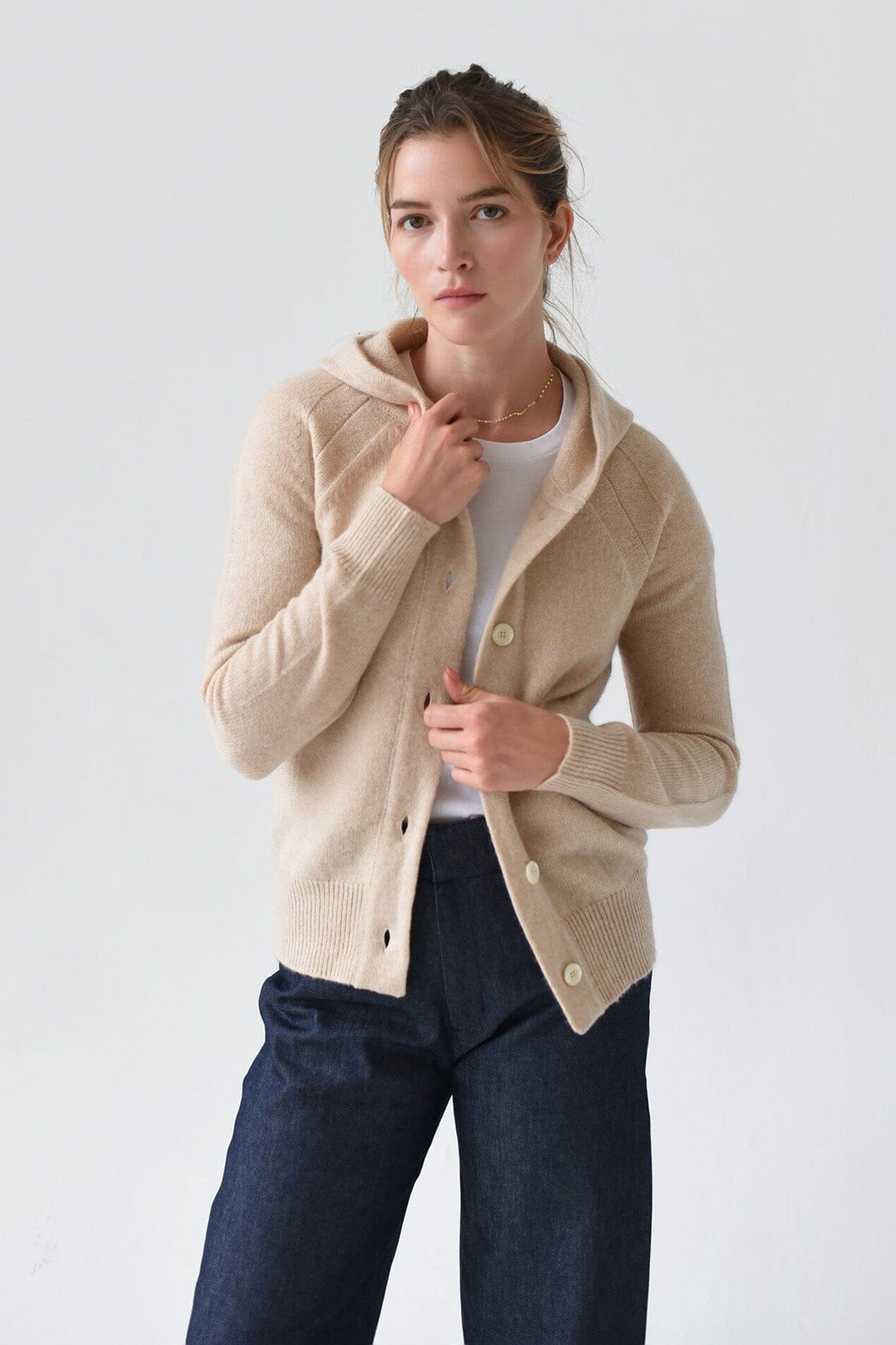 A woman wearing a beige cashmere hoodie with mother of pearl buttons and ribbed trims, paired with a white top and blue jeans