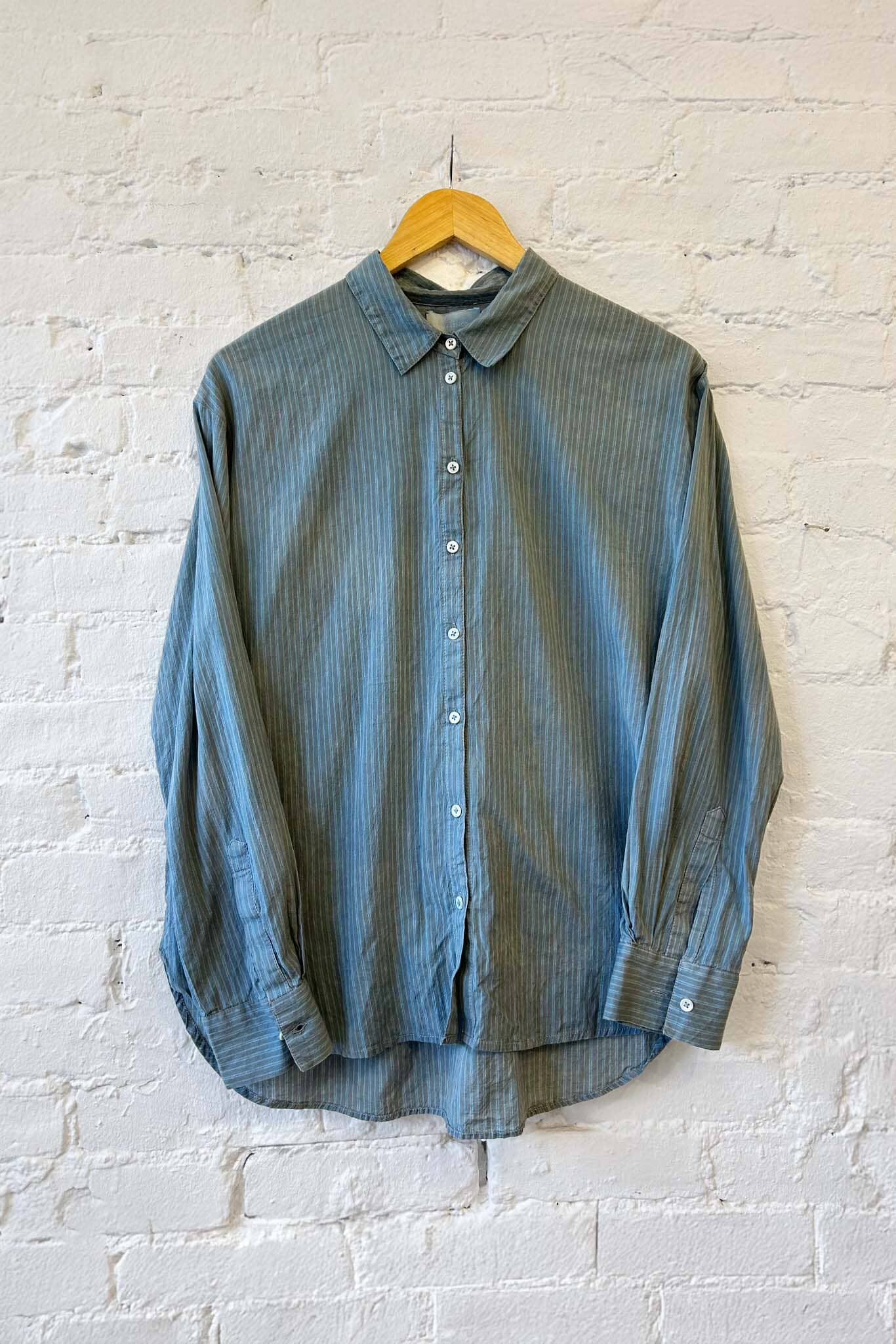 Blue green striped long sleeve cotton shirt on a hanger against a white brick wall