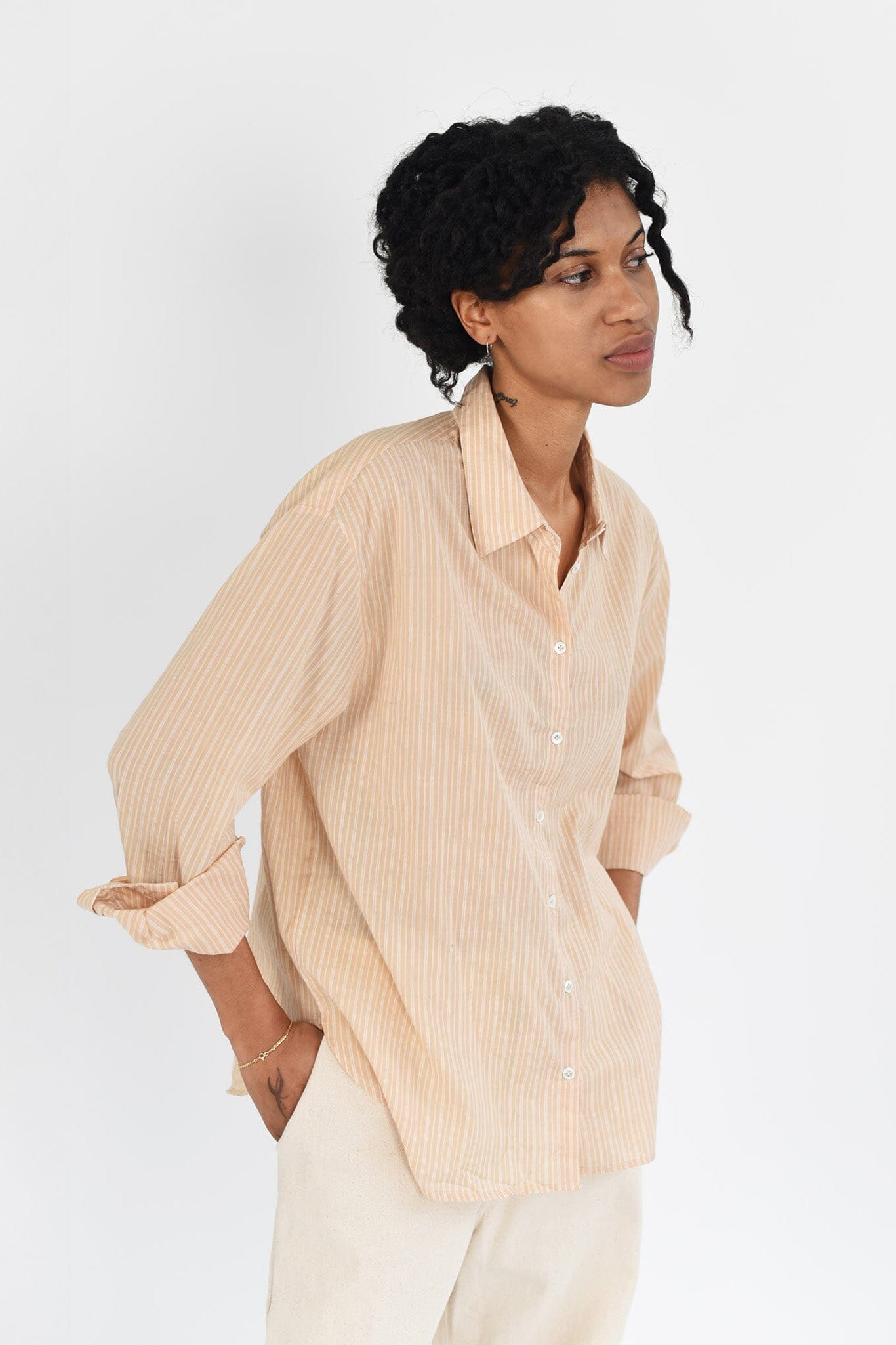 A woman wears a tan, golden striped long sleeve button down blouse. The top is lightweight and made of cotton. Available to purchase at M.Patmos in Brooklyn, New York. 