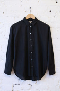 A black long-sleeve button-up shirt hanging on a wooden hanger against a white painted brick wall.