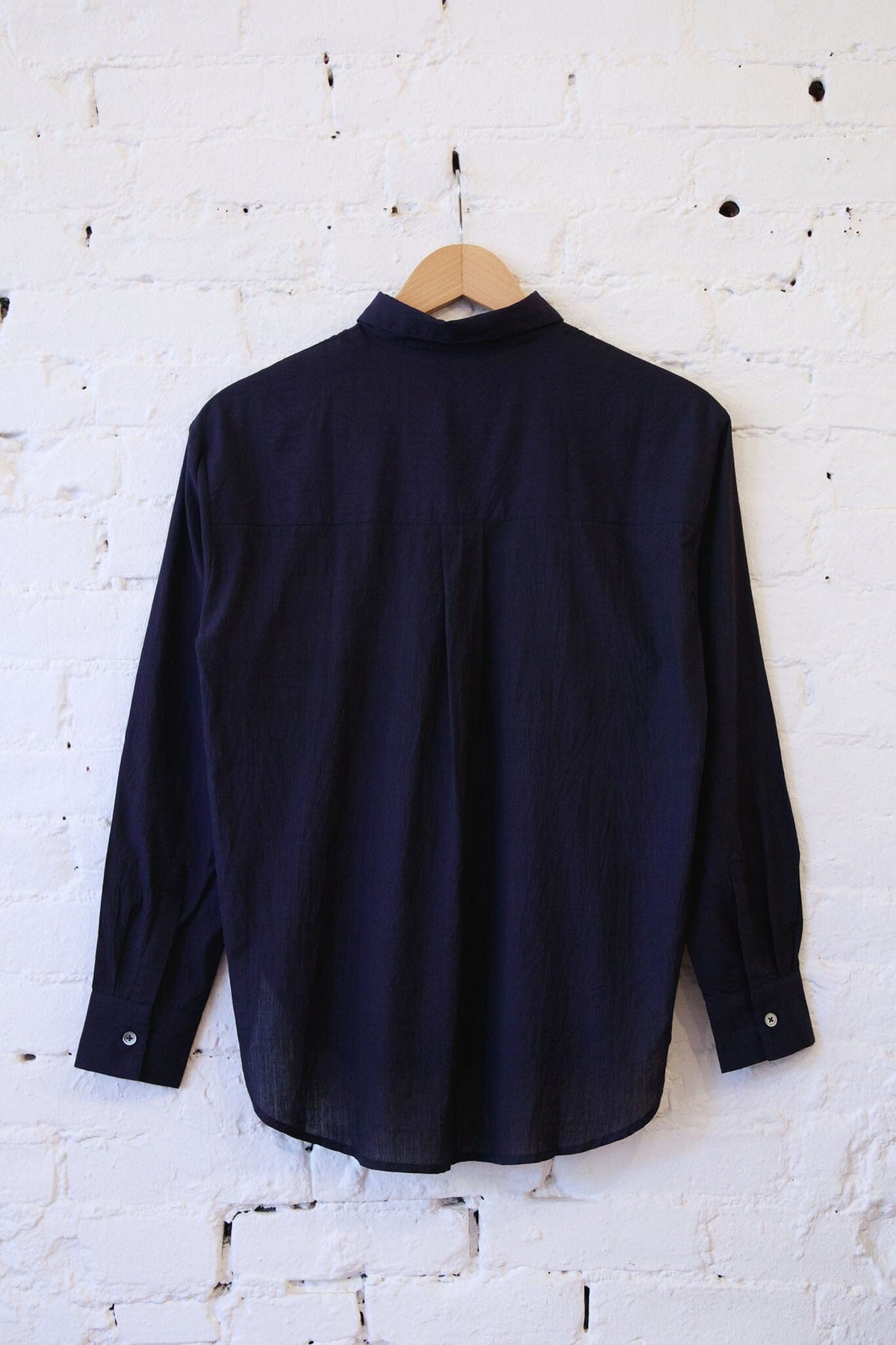A dark navy blue long-sleeve shirt hanging on a wooden hanger against a white painted brick wall.
