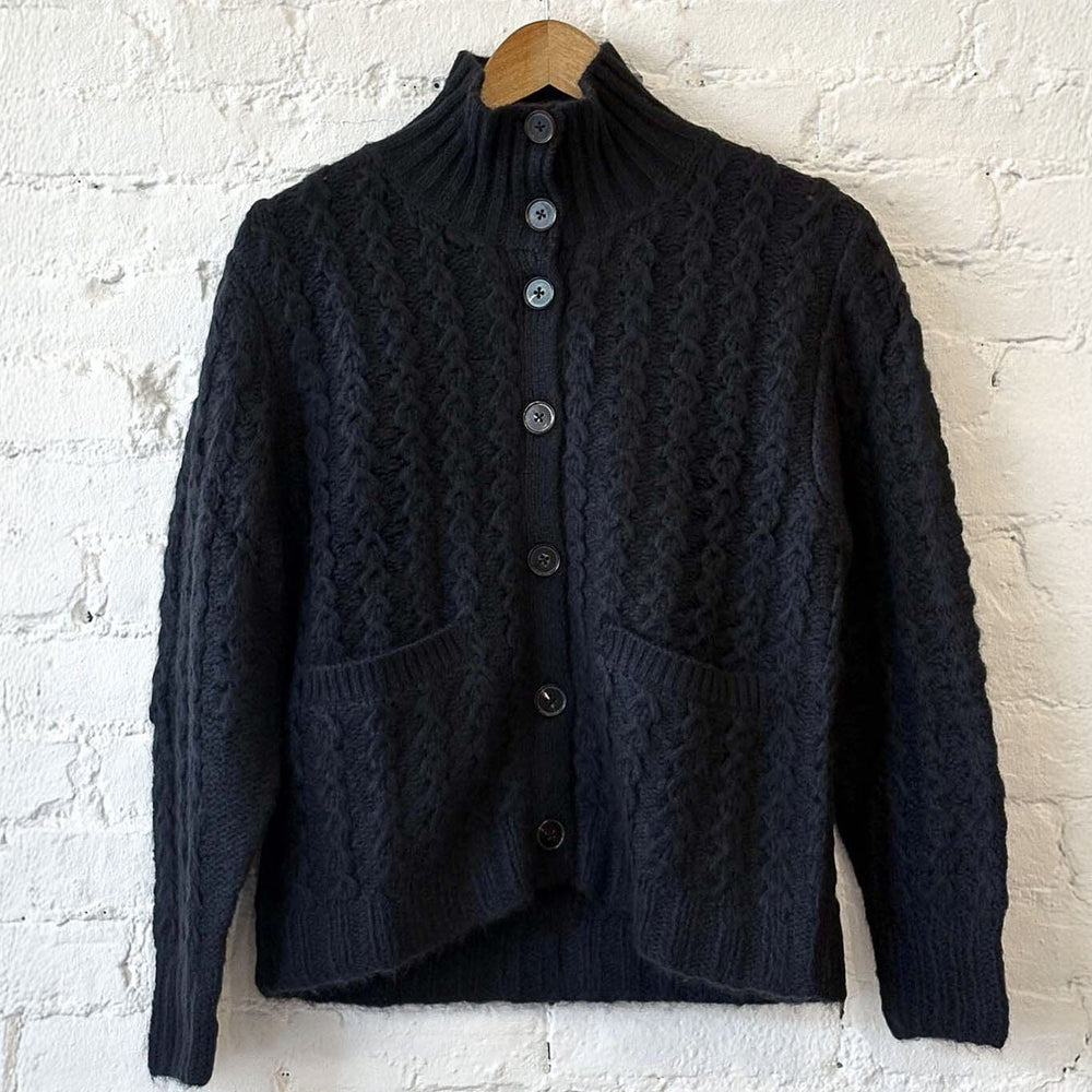 
                  
                    Black knitted cardigan on a wooden hanger against a white brick wall.
                  
                
