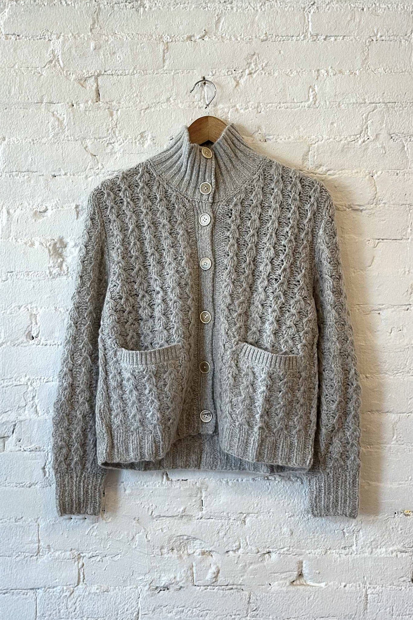 Gray knitted cardigan on a wooden hanger against a white brick wall.