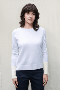 A woman with shoulder-length brown hair wearing an icy blue crewneck sweater with contrasting cream cuffs and dark navy pants, standing against a light beige textured wall.