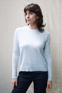 A woman wearing a light blue knit crewneck sweater and dark navy pants, standing against a neutral beige fabric backdrop.