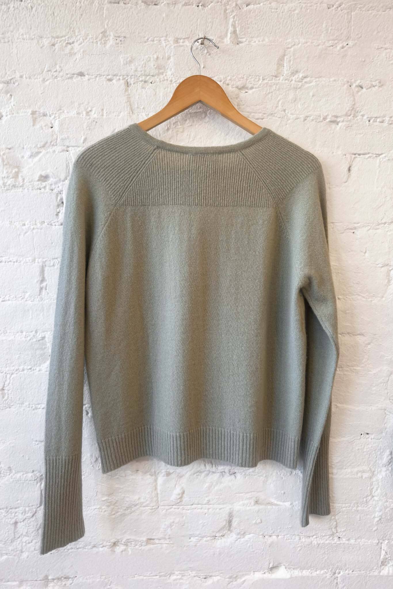Sage green sweater on a wooden hanger against a white brick wall.