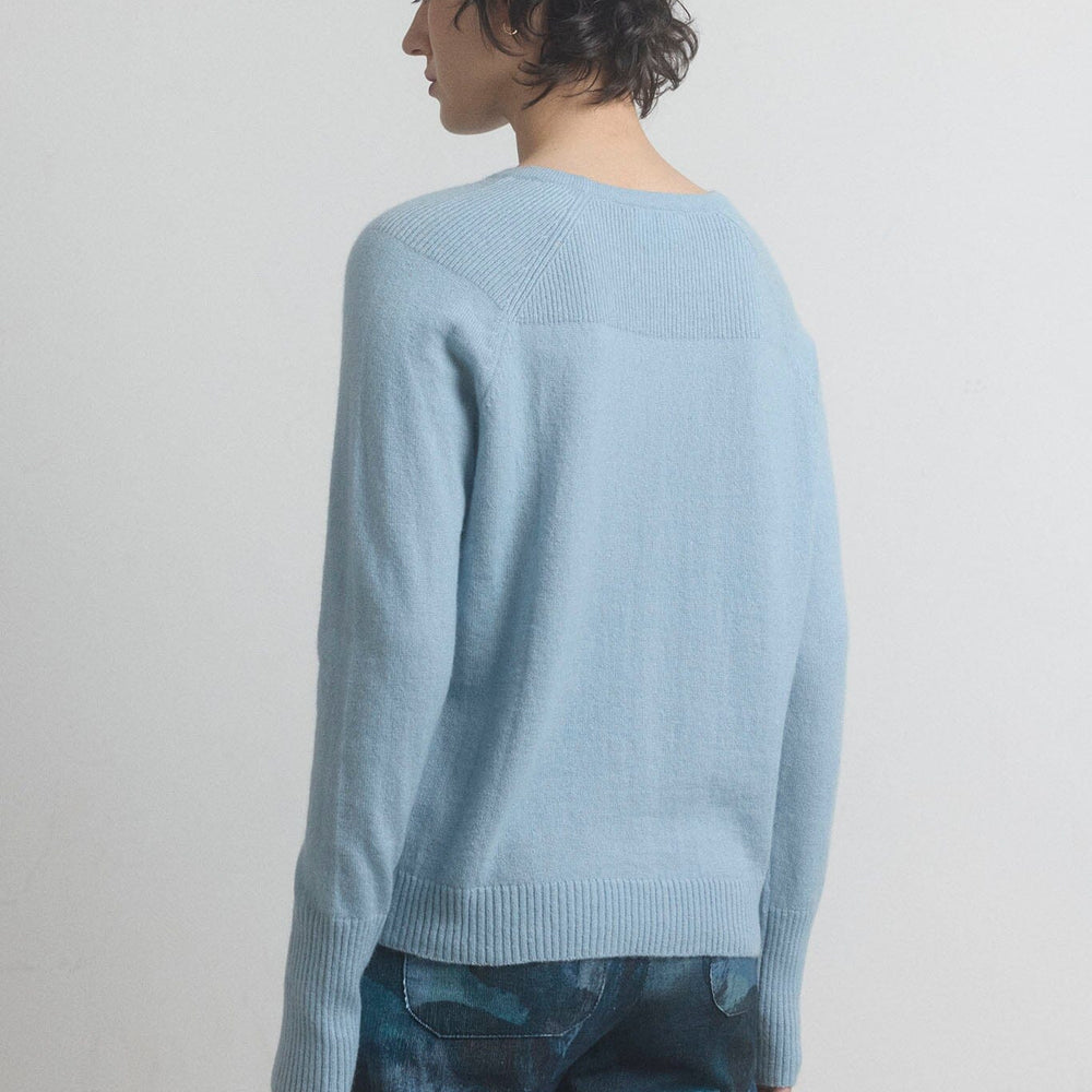 The back view of a woman wearing a light blue sweater and patterned pants on a plain background