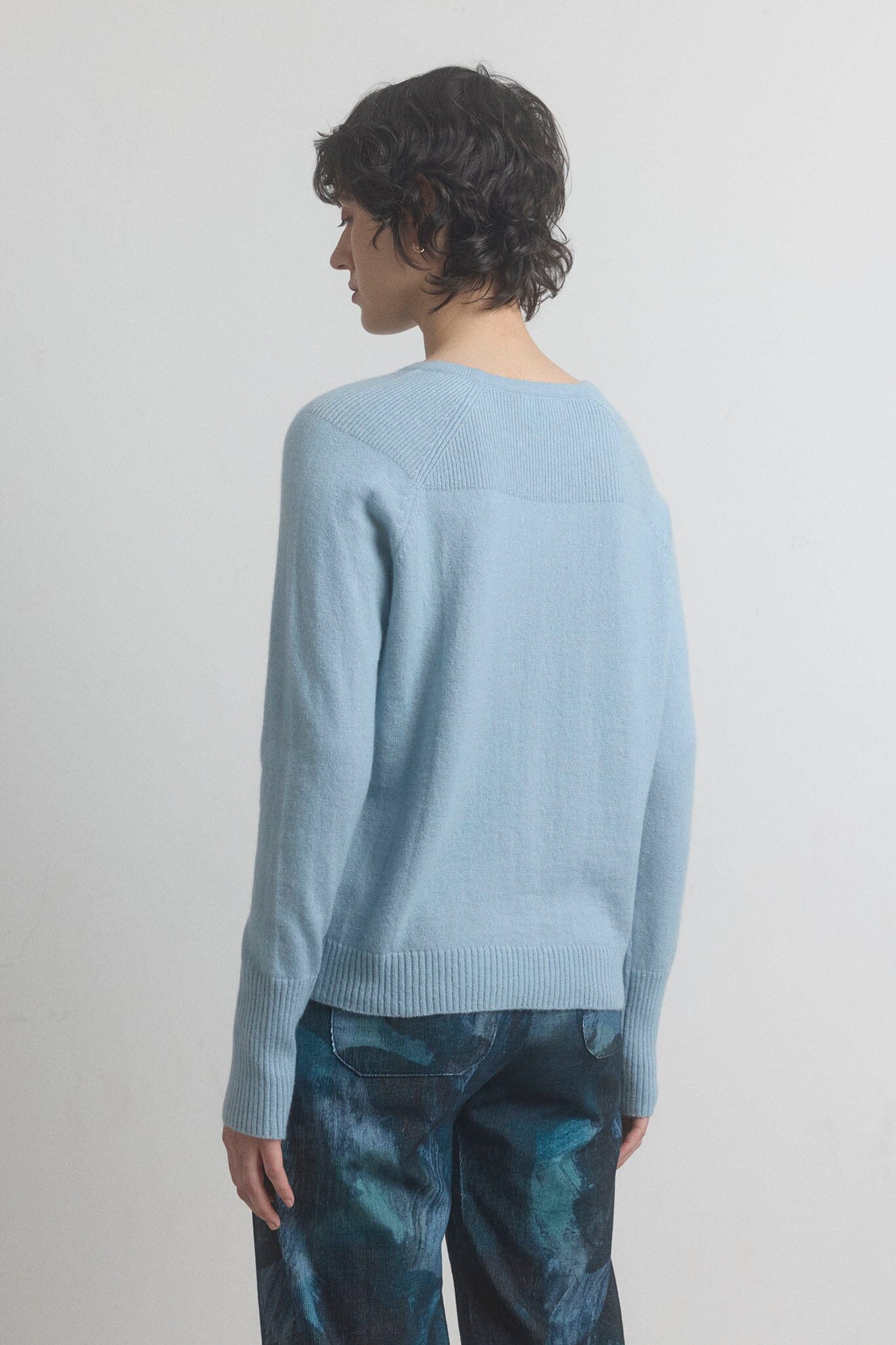 The back view of a woman wearing a light blue sweater and patterned pants on a plain background