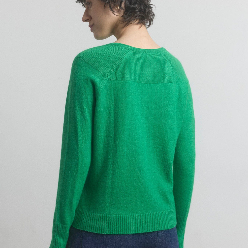 Woman facing the back wearing a green sweater and blue pants against a plain background