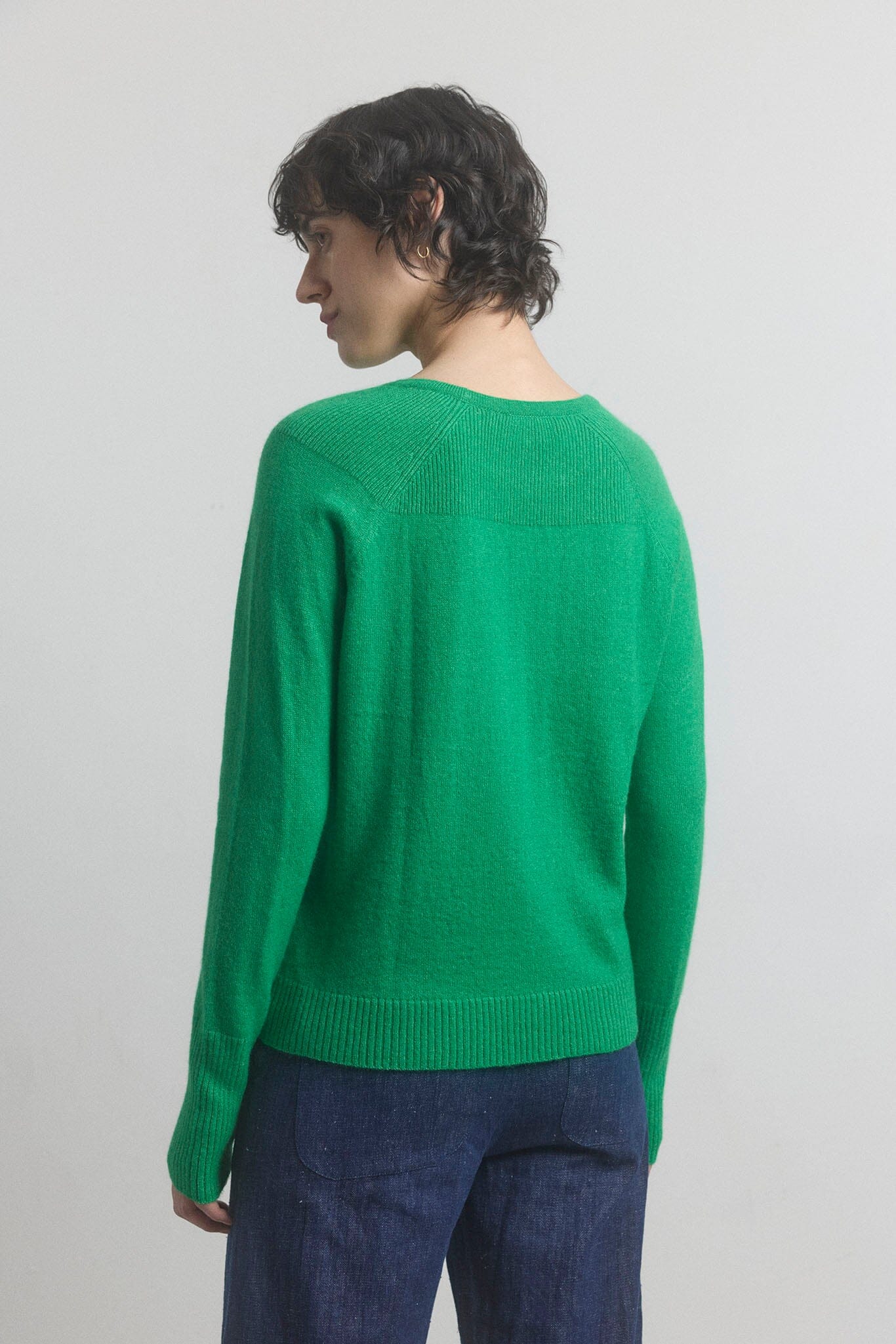 Woman facing the back wearing a green sweater and blue pants against a plain background